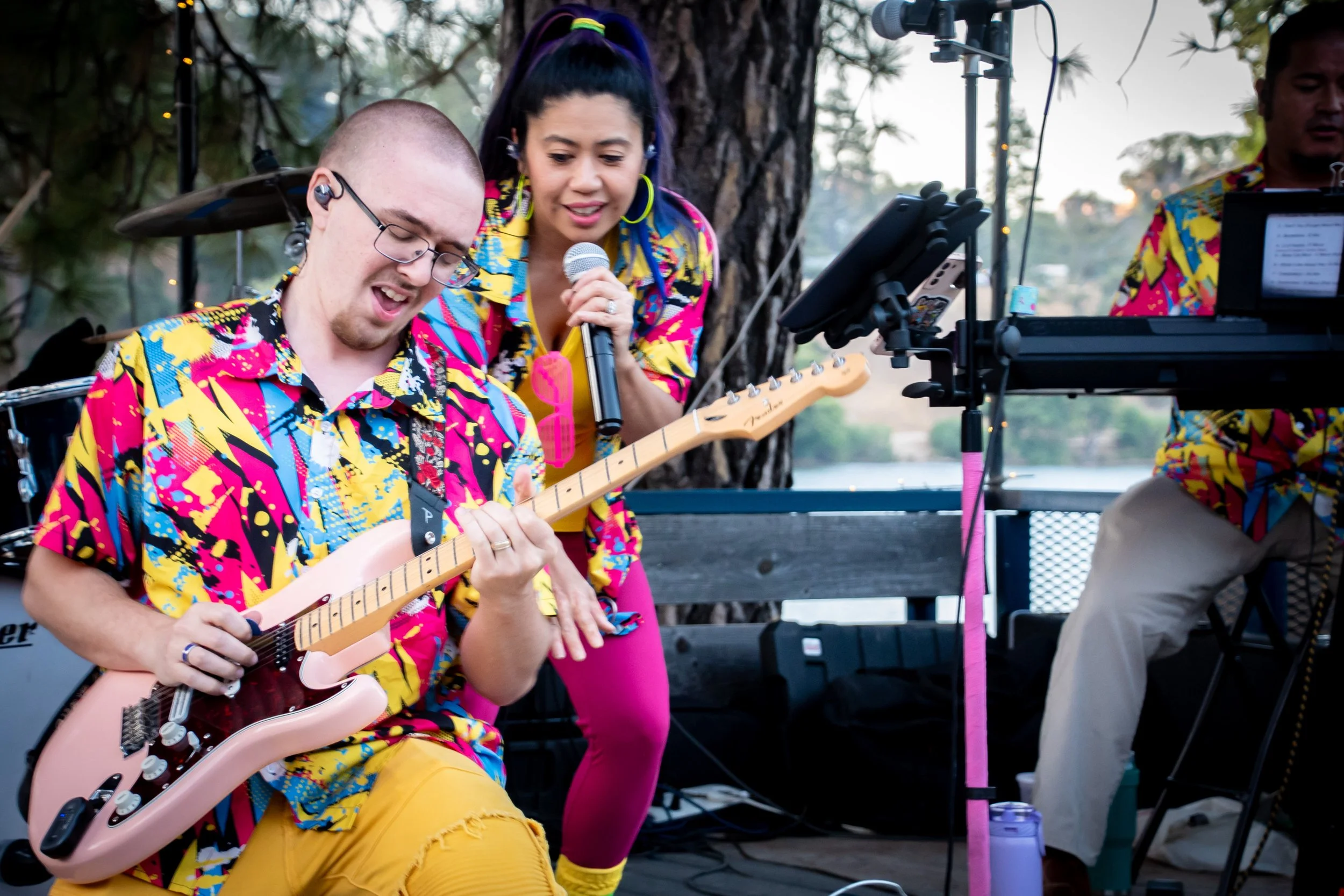 Three musicians performing outdoors, wearing colorful, patterned shirts, with one playing an electric guitar and another singing into a microphone, surrounded by musical equipment, trees, and a lake in the background.