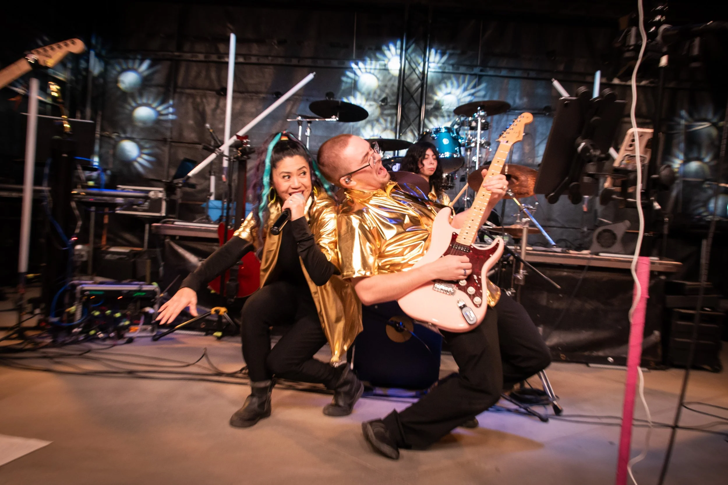 Three musicians performing on stage with musical instruments, colorful lighting, and stage equipment.