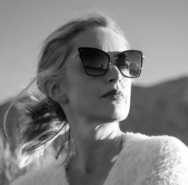 Black and white photo of a woman, Claire Von Cleveland, with blonde hair wearing large sunglasses, outdoors with mountains in the background.