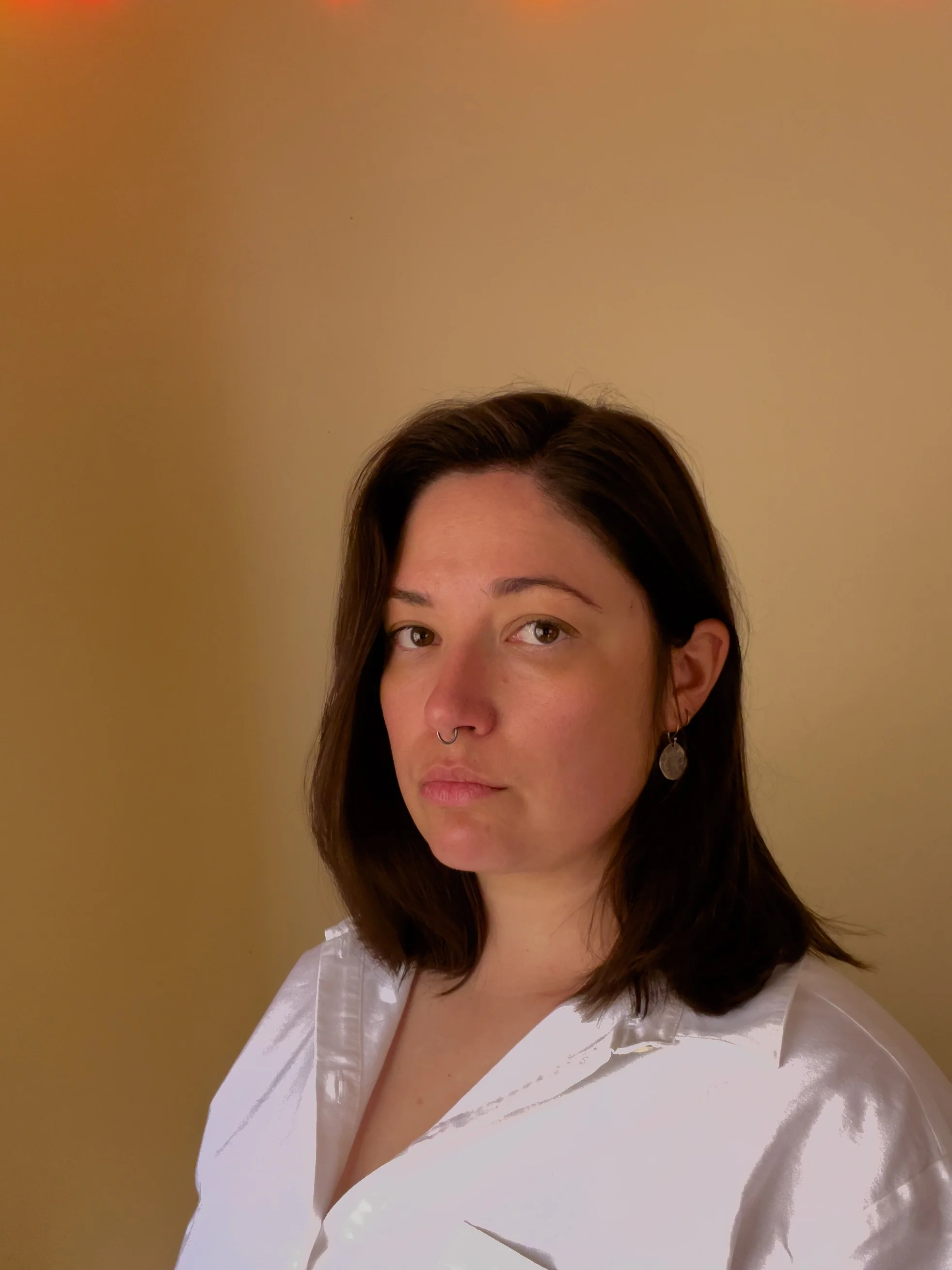 A woman with shoulder-length dark hair, wearing a white satin blouse and earrings, standing against a plain beige wall.