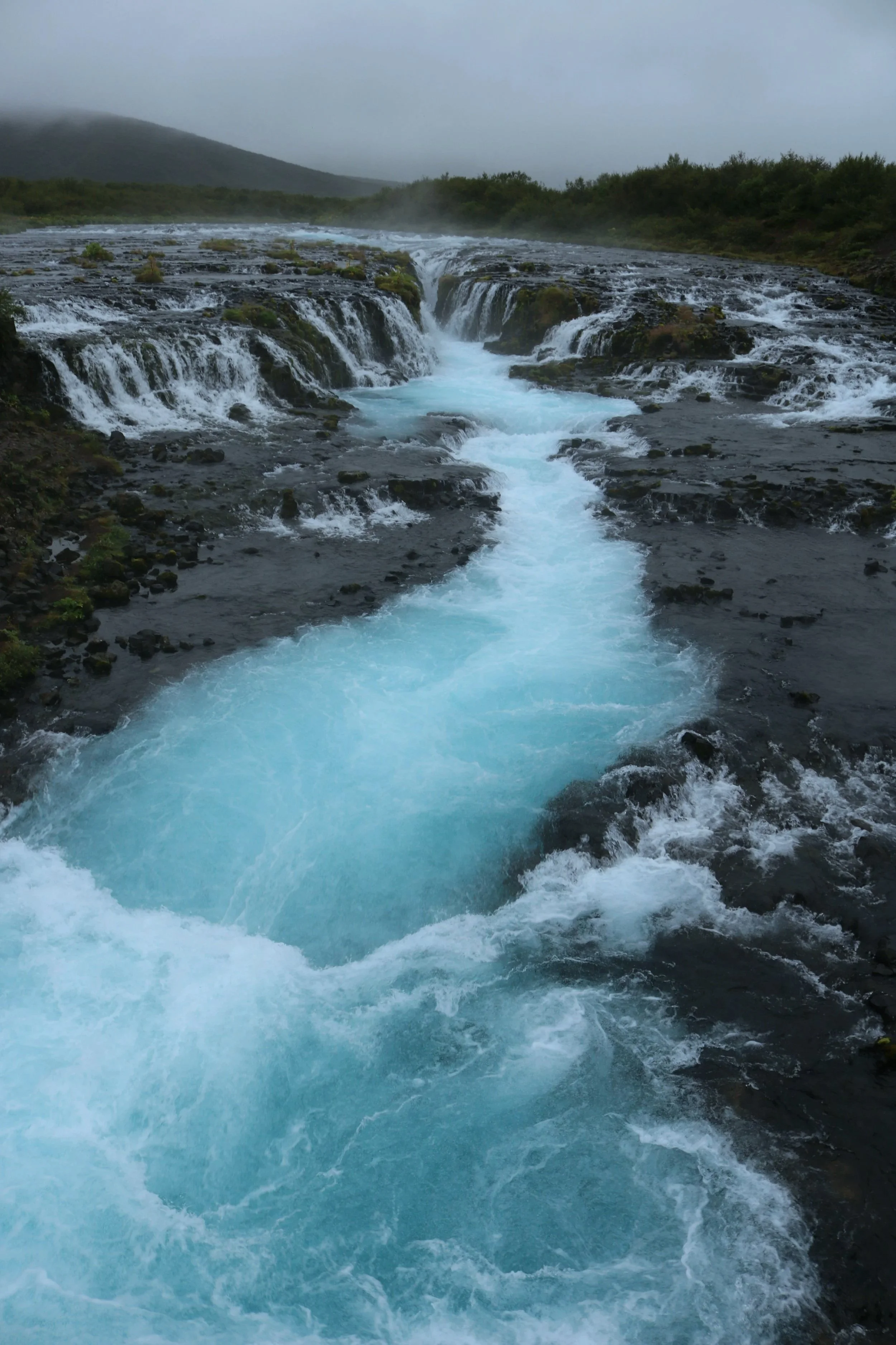 A river with blue water flows over rocks and small waterfalls in a lush, green landscape with mountains in the background under a cloudy sky.
