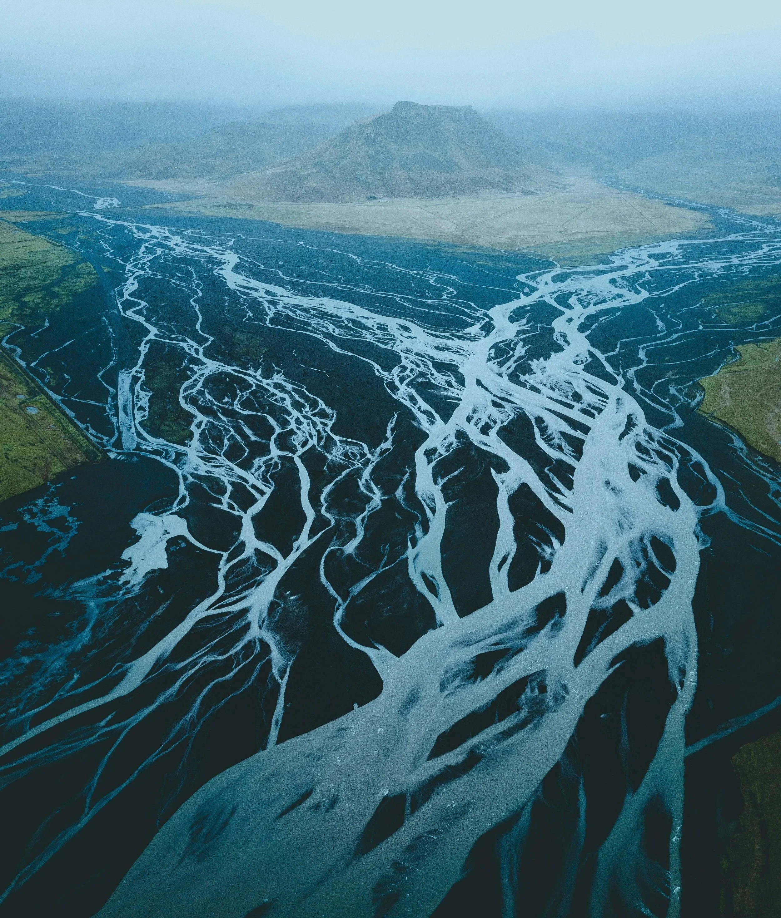 Aerial view of a river delta with numerous braided channels flowing into a lake, with a mountain in the background under a cloudy sky.