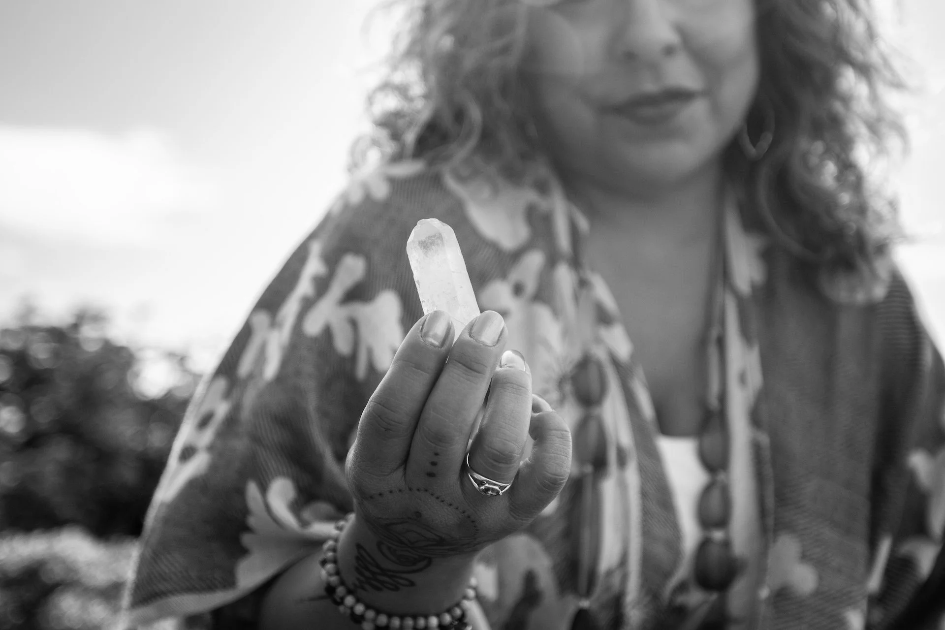 A woman with curly hair, wearing a patterned shirt, holding a clear crystal wand close to the camera. Her face is partially visible, and she has tattoos and jewelry on her hand and wrist. The background is blurred outdoors.