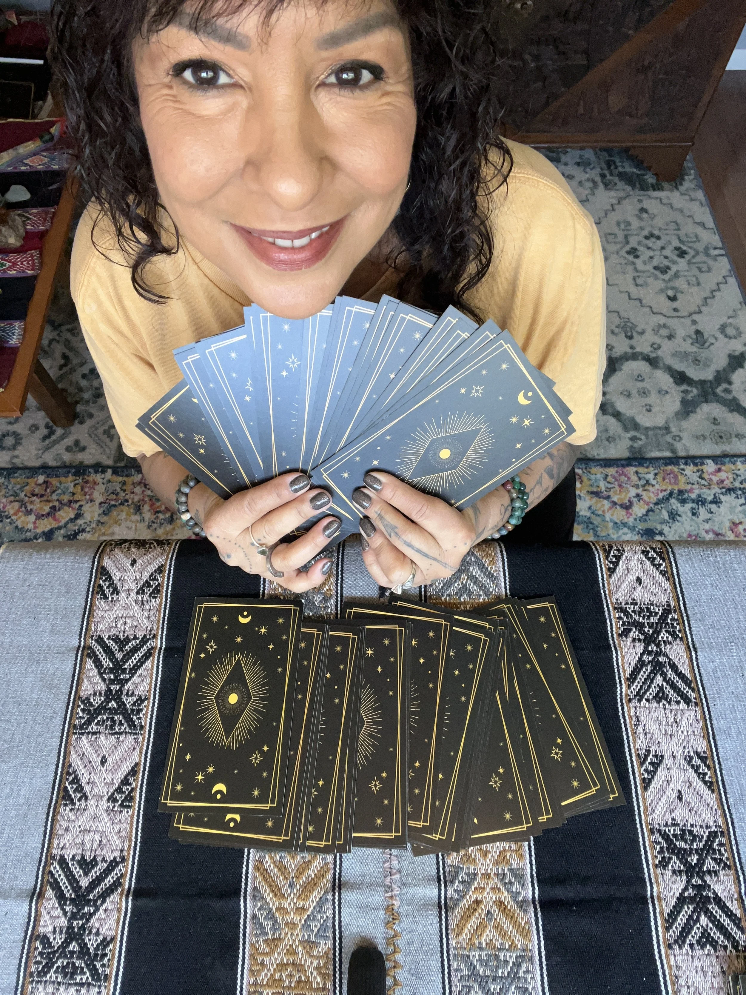 A woman with dark curly hair smiling and holding tarot cards, with more tarot cards spread out on a table in front of her, in a room with a patterned rug and wooden furniture.