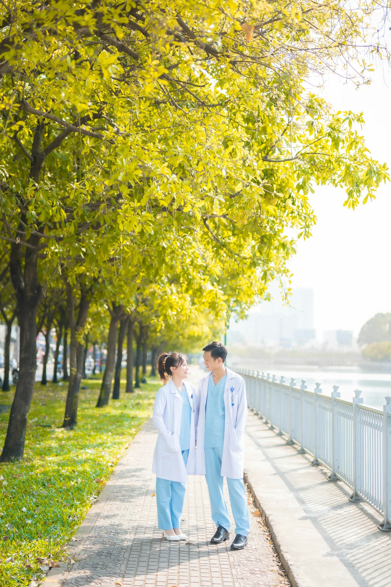 Two healthcare professionals, a woman and a man, walking and talking along a pathway beside a river, under a canopy of green and yellow autumn trees, with sunlight filtering through.