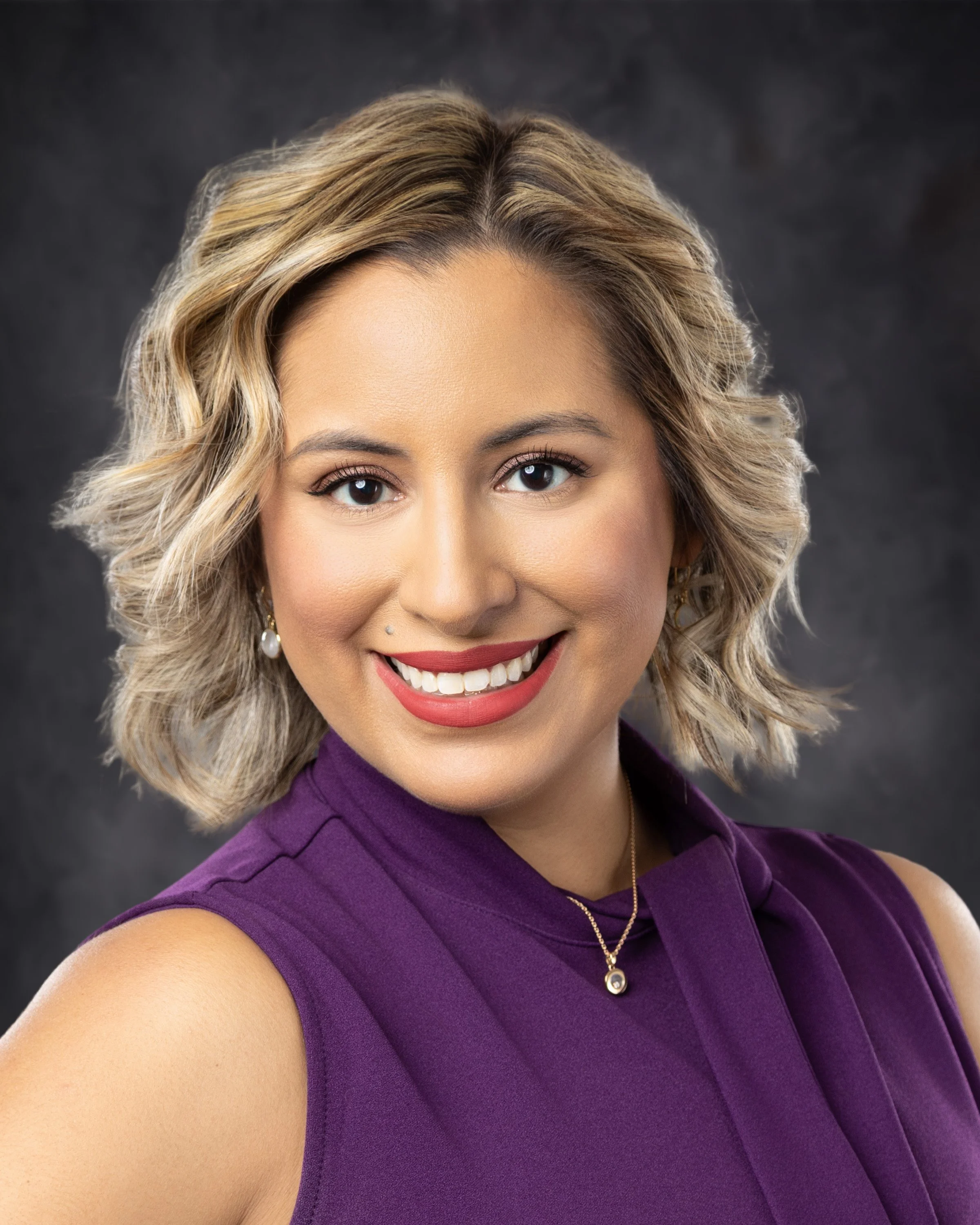 A smiling woman with blonde wavy hair, wearing a purple sleeveless top, gold earrings, a small pendant necklace, and makeup, posed against a dark gray background.