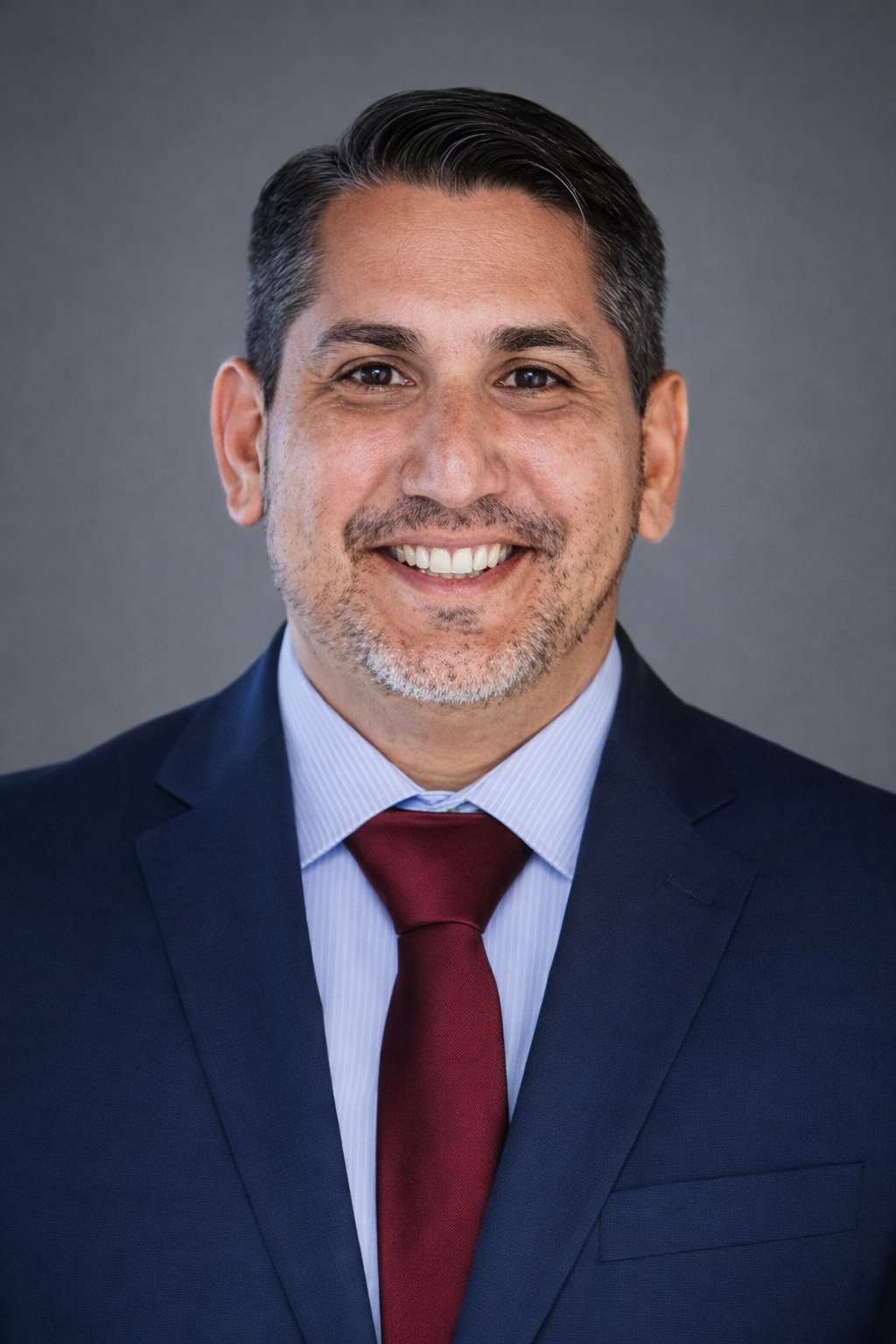 Professional headshot of a smiling man with dark hair, beard, and mustache, wearing a navy suit, light blue dress shirt, and maroon tie, against a plain gray background.