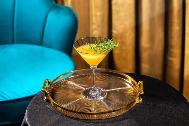 A yellow cocktail garnished with rosemary sprigs on a glass tray on a black table, with a blue velvet armchair and wooden curtains in the background.