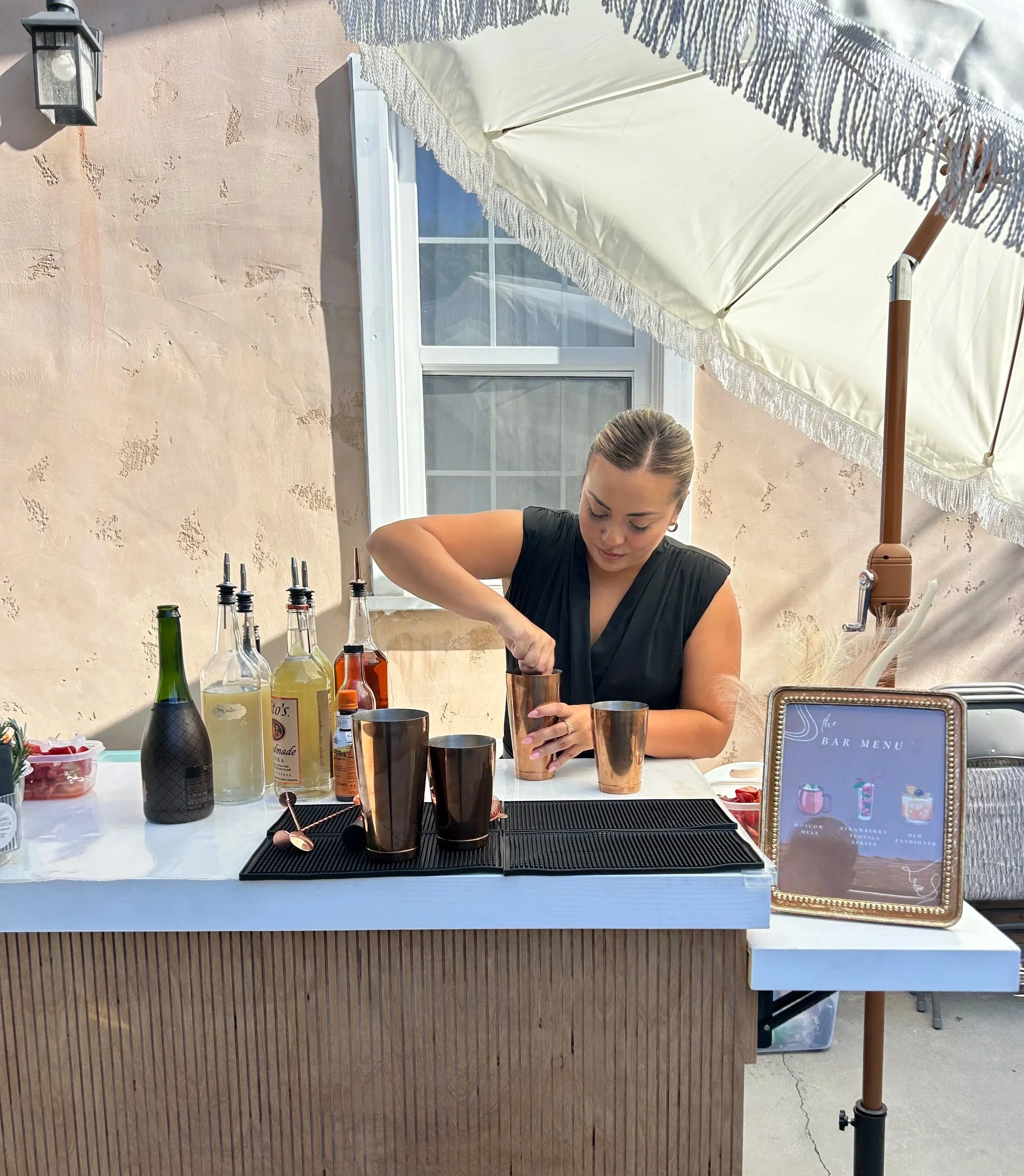A woman preparing drinks at an outdoor bar under a large white umbrella, with liquor bottles on the counter and a framed bar menu sign nearby.