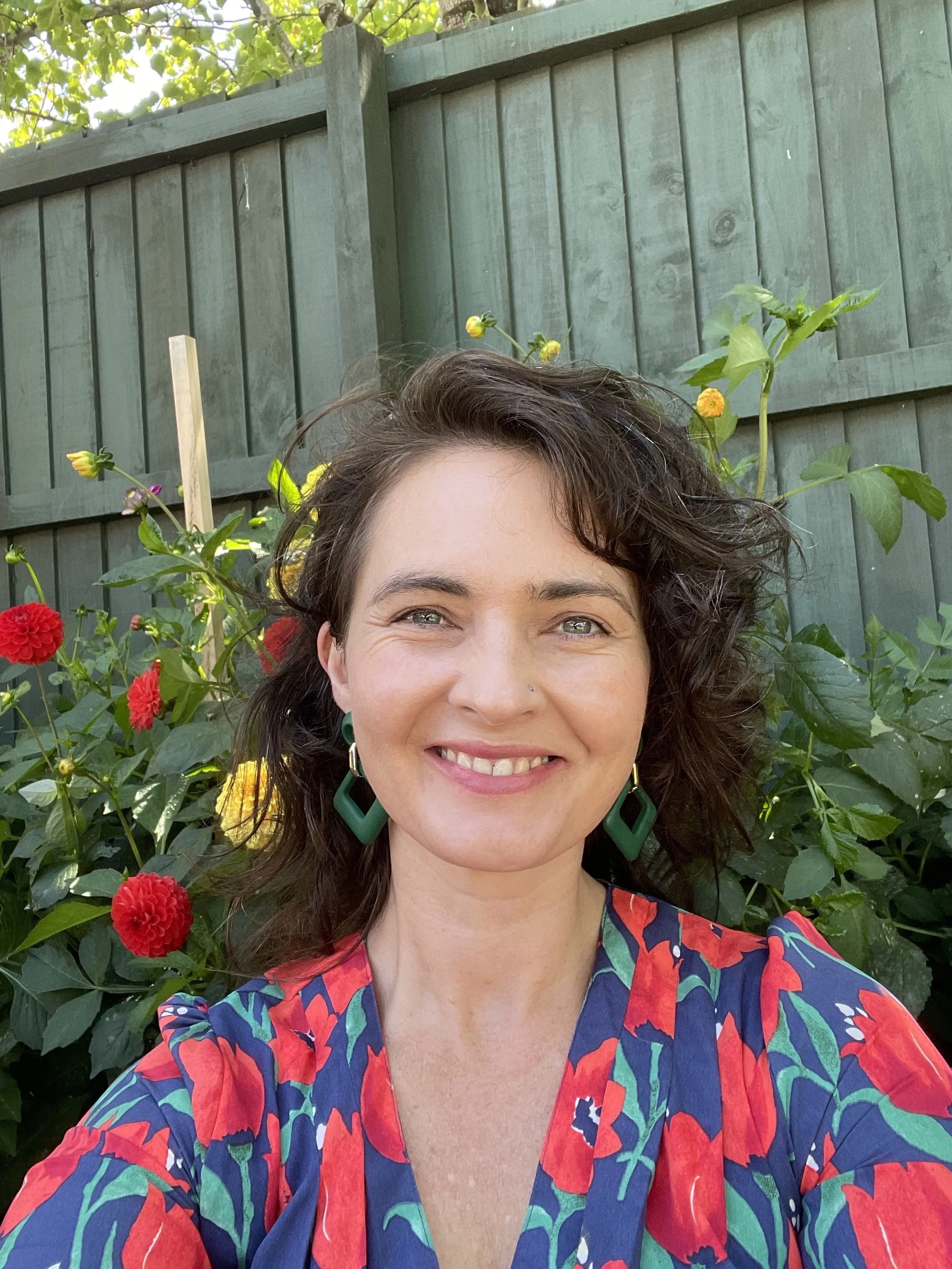 A woman smiling outdoors in front of green plants and flowers, wearing a colorful floral blouse and earrings.