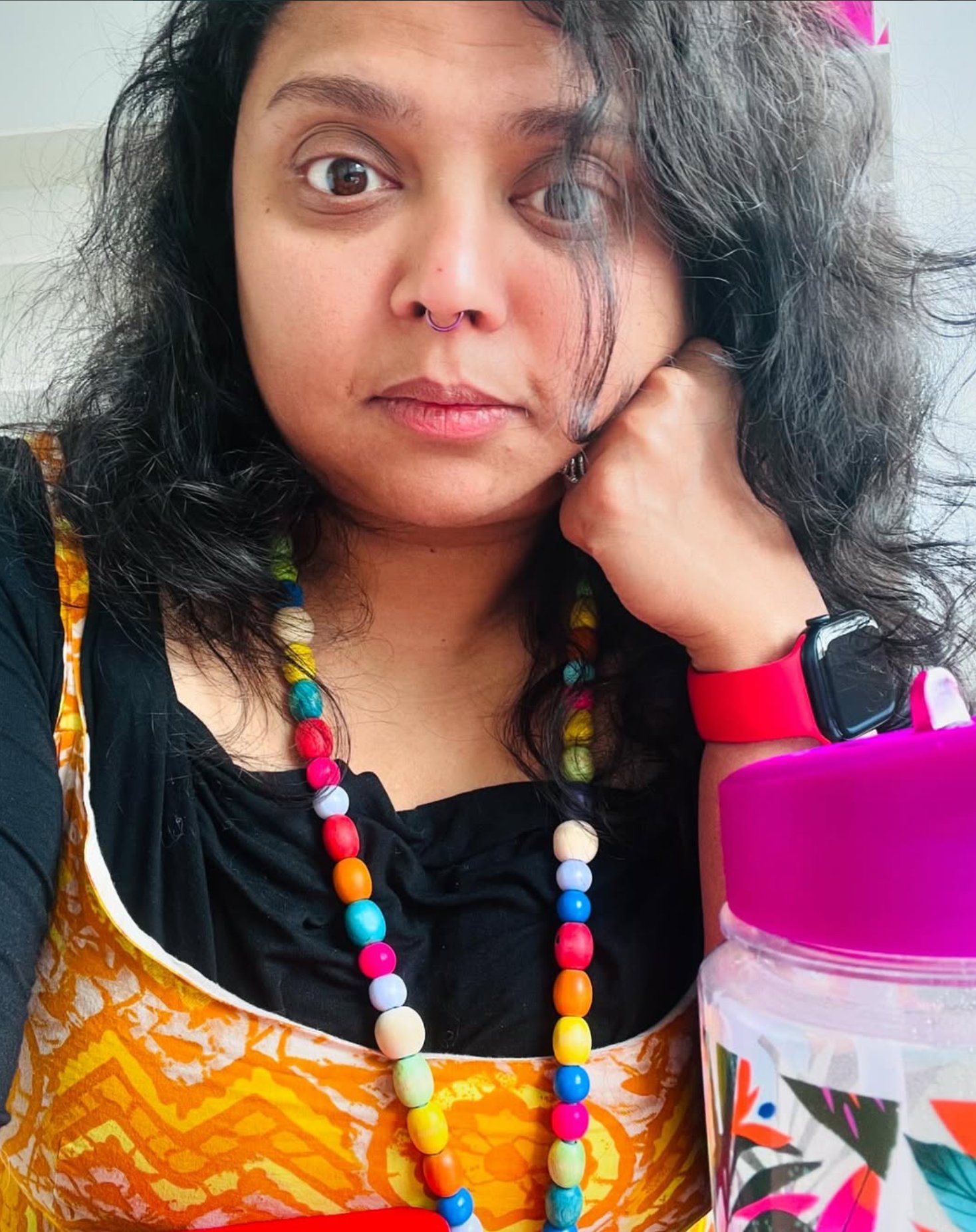 A woman with curly dark hair, wearing a black top, colorful beaded necklace, a colorful apron, a pink wristwatch, and a colorful water bottle, sitting indoors and resting her face on her hand.