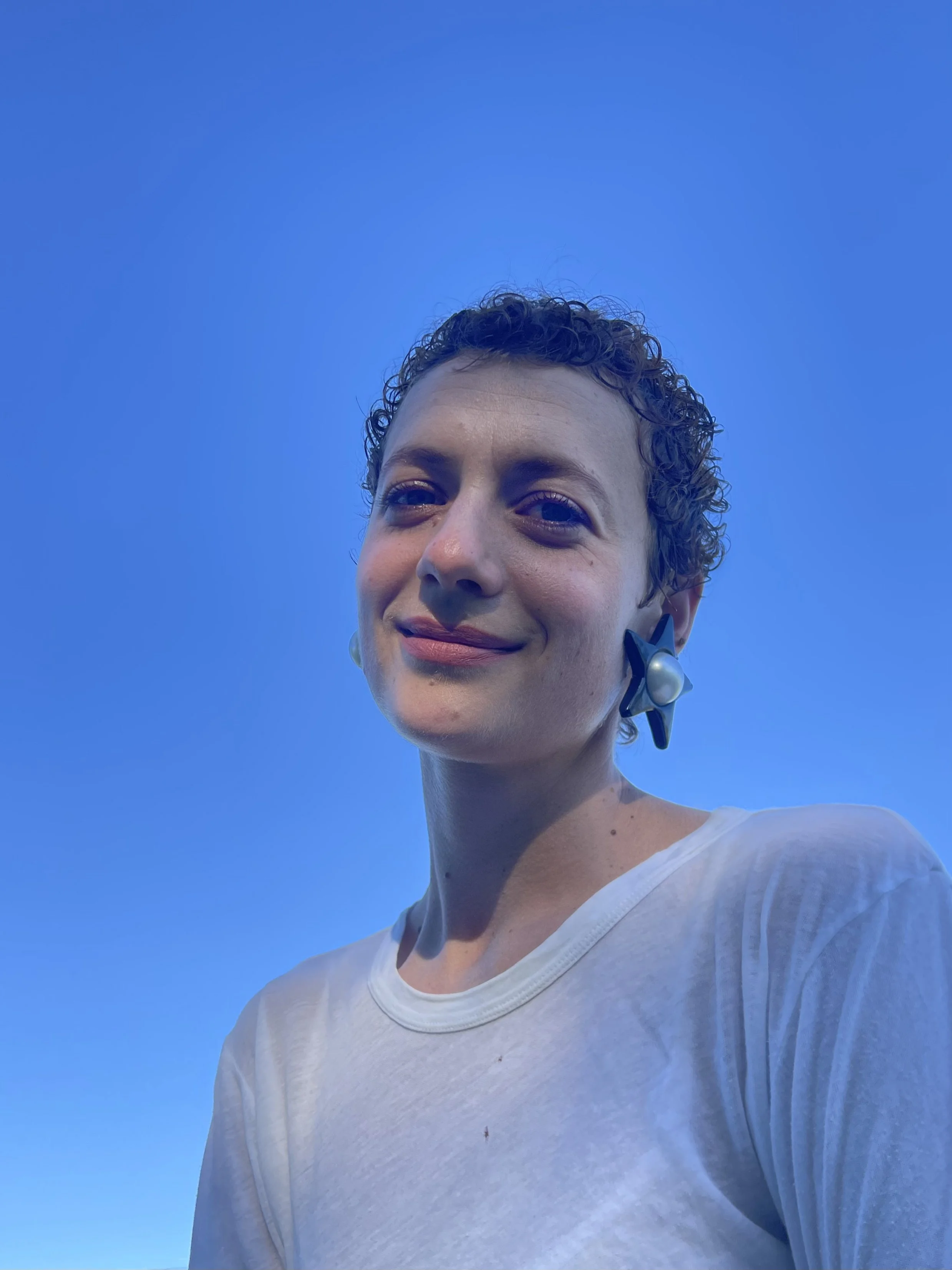 A woman with short, curly hair wearing star-shaped earrings and a white shirt, standing against a clear blue sky.
