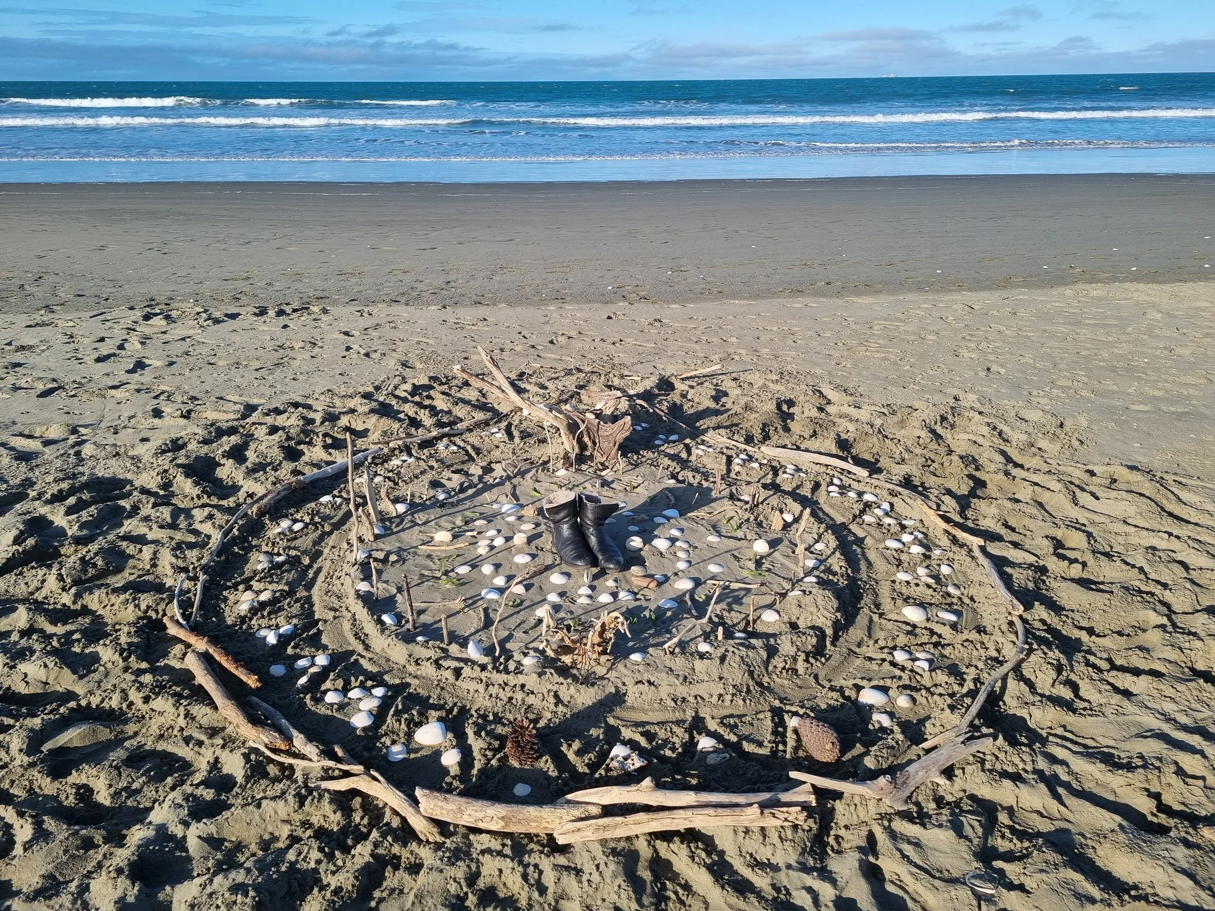 A sandy beach with a makeshift memorial circle made of driftwood, shells, and other natural materials. In the center, there is a pair of black shoes. The ocean and sky are visible in the background.