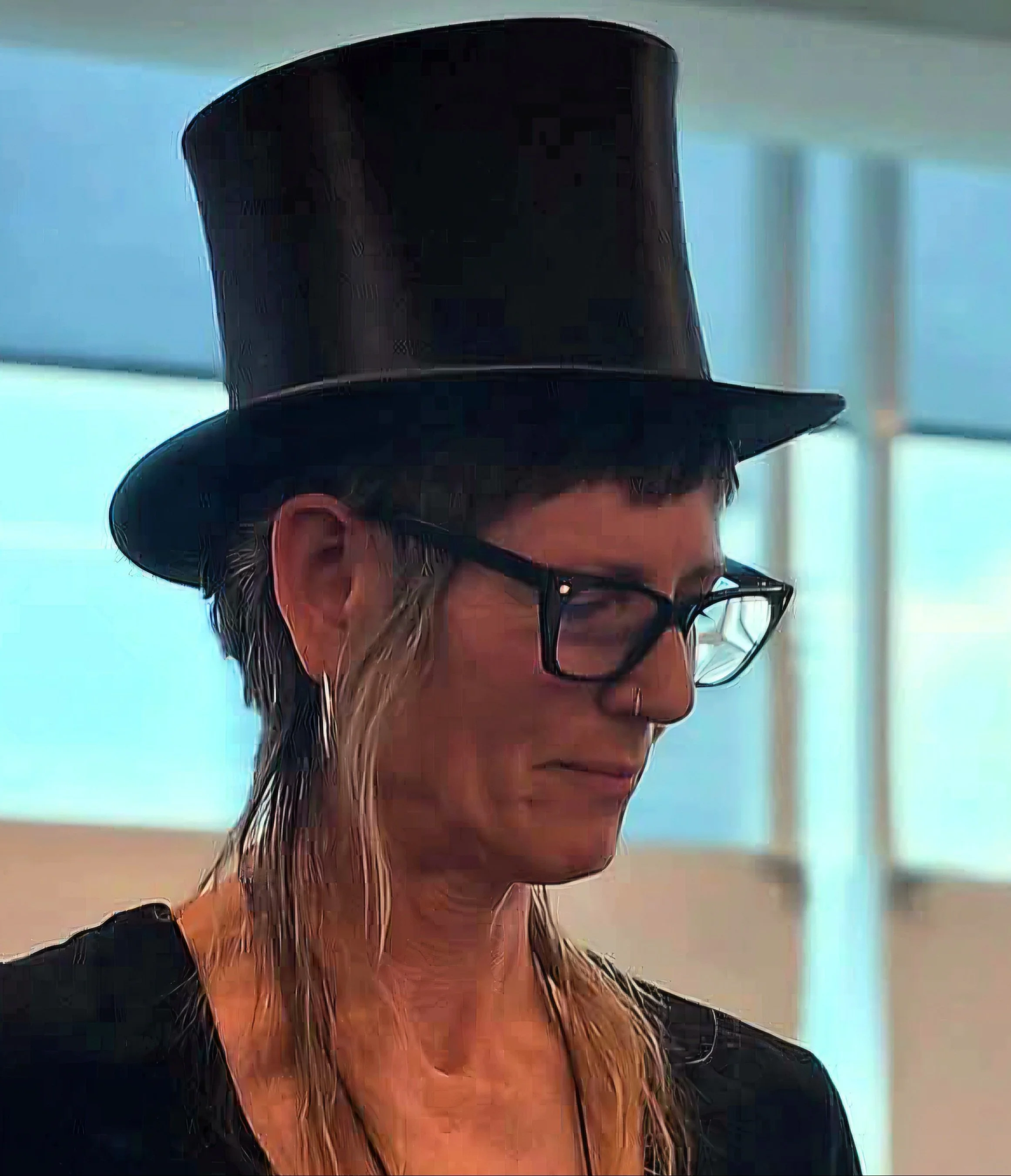 A woman wearing a black top hat, glasses, and a nose ring, standing indoors near a window.