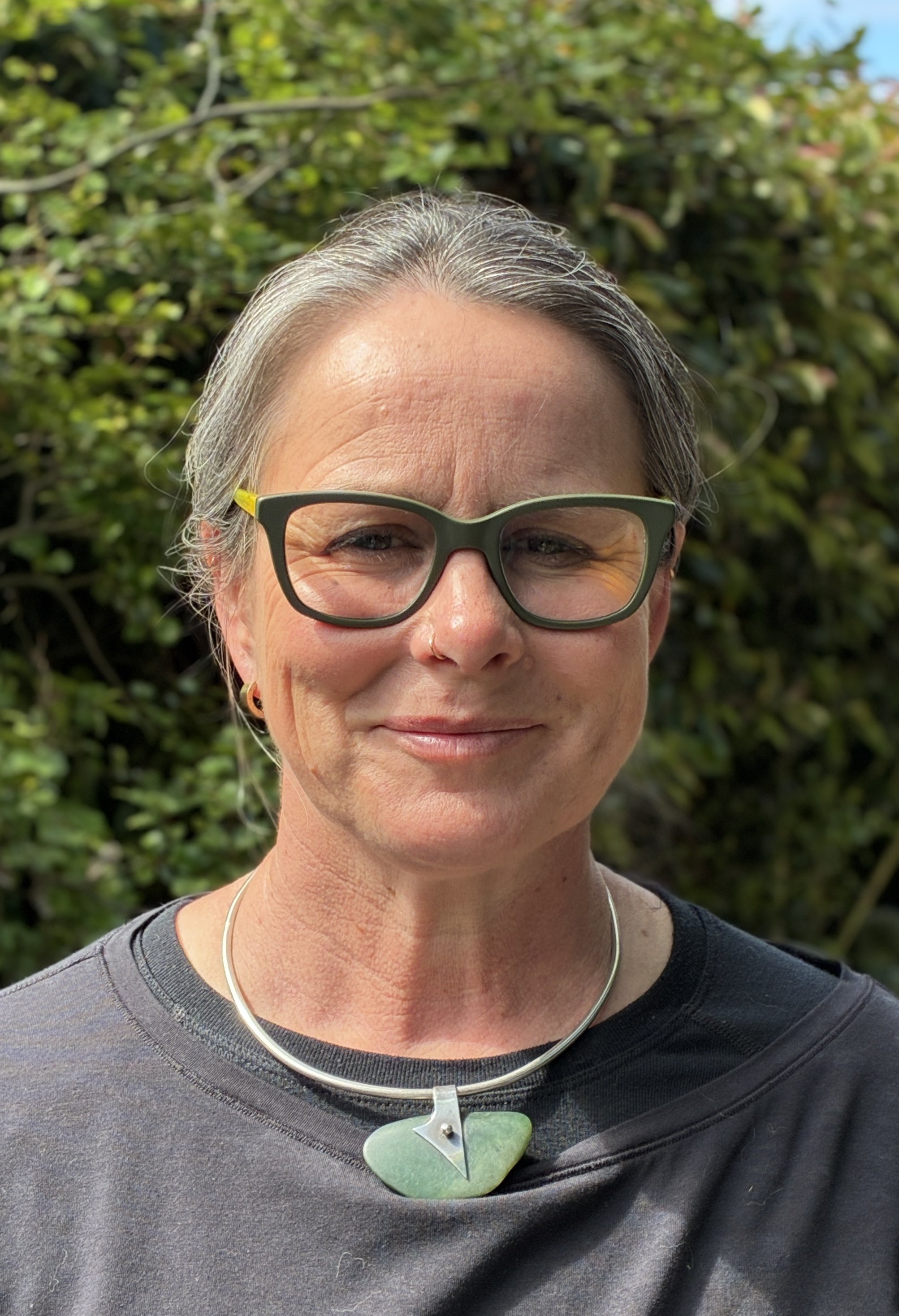 A middle-aged woman with gray hair, wearing green glasses, smiling outdoors in front of green foliage, with a black shirt and a large green and silver necklace.