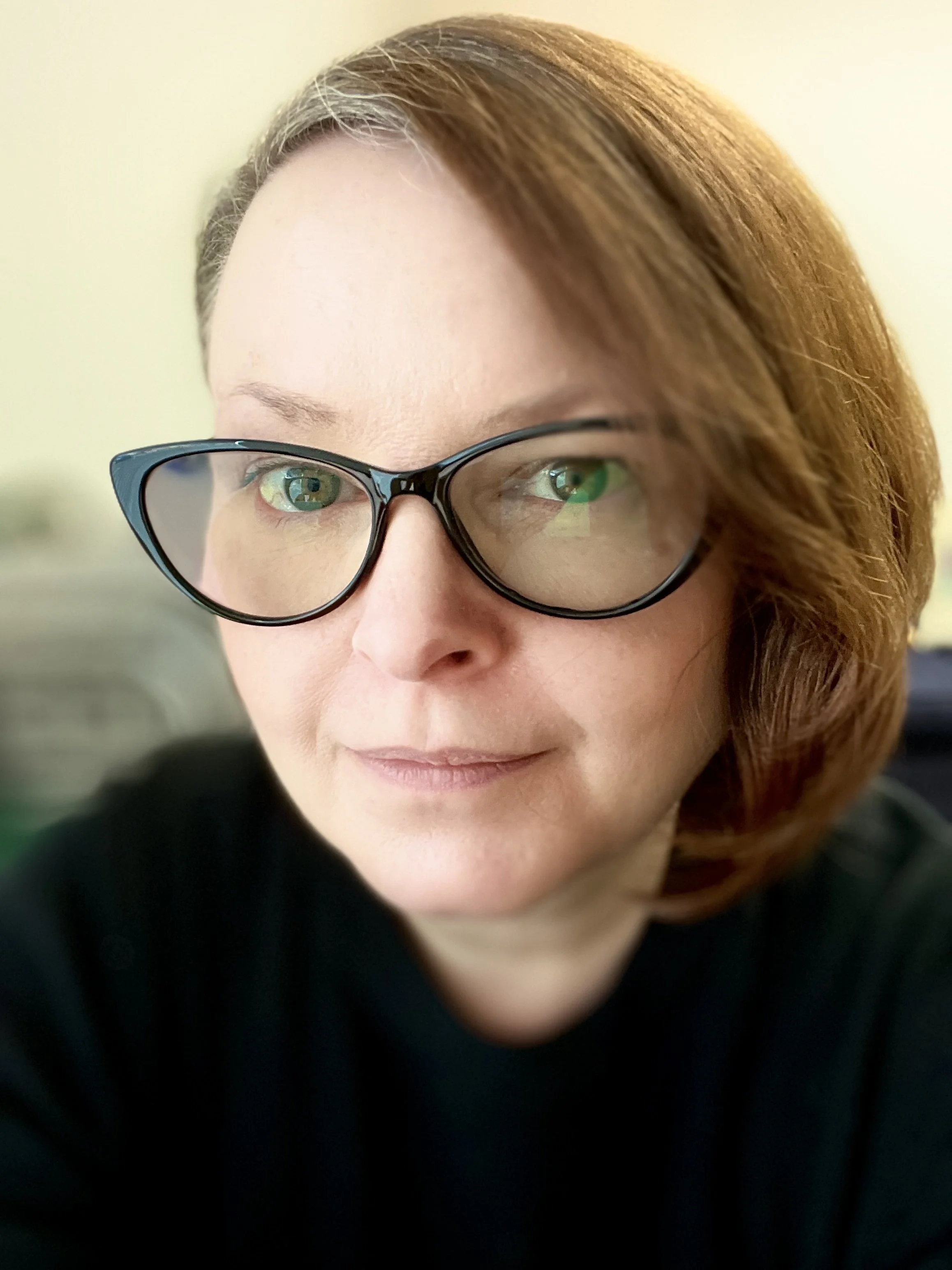 A woman with short reddish-brown hair and glasses, looking at the camera with a slight smile in an indoor setting.