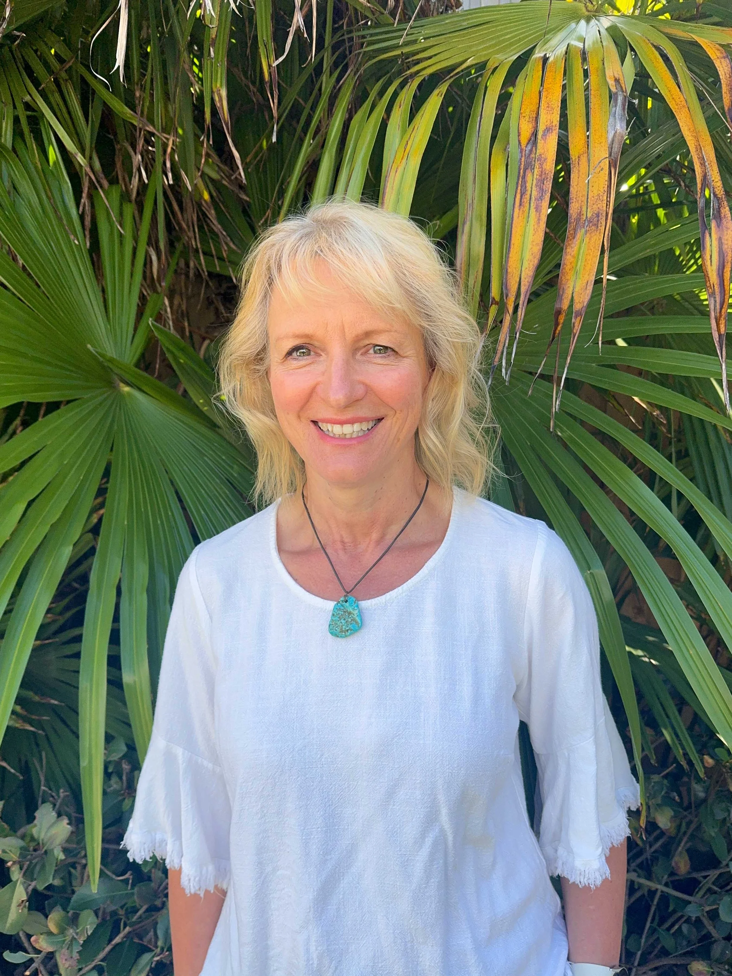 A smiling woman with blonde hair and a turquoise pendant necklace standing in front of lush green tropical plants.