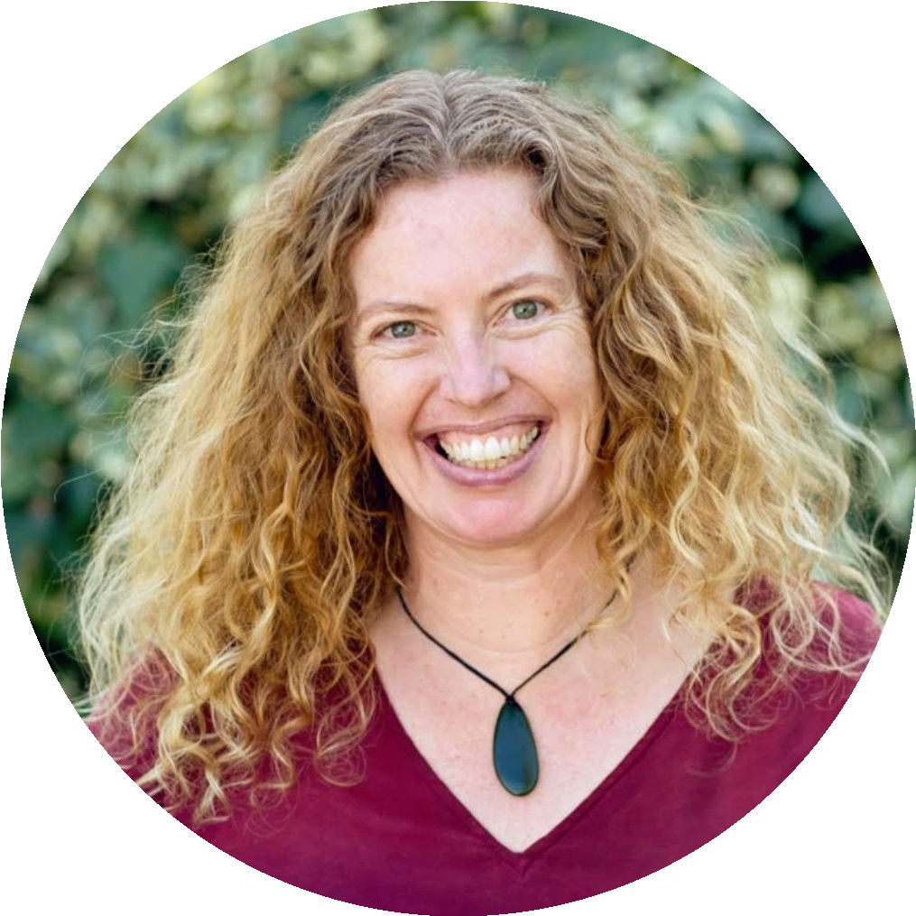 A smiling woman with curly red hair wearing a maroon top and a black pendant necklace standing outdoors with green foliage in the background.