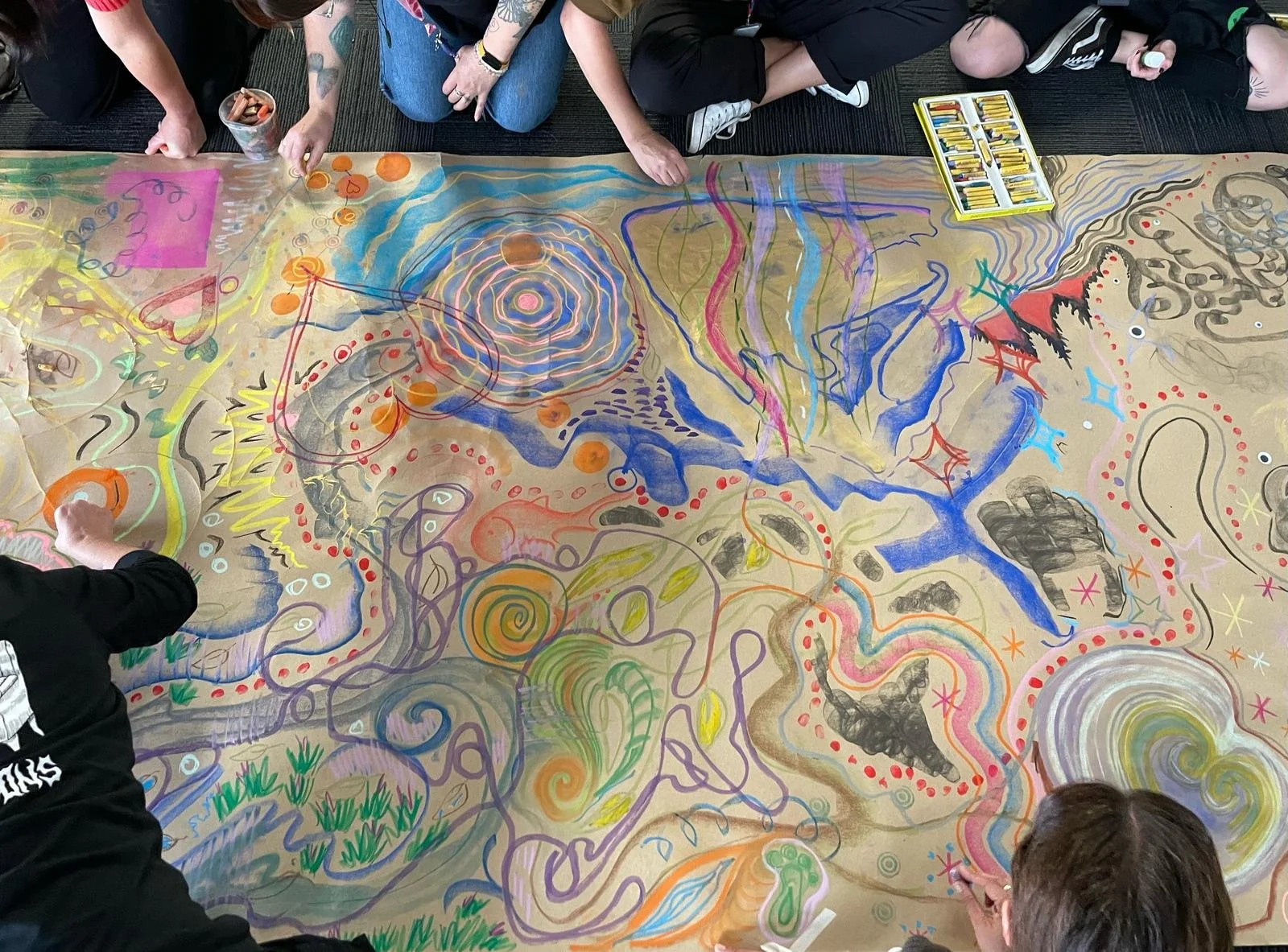 Colorful abstract artwork on a large sheet of paper with people working on it, using paints and markers.