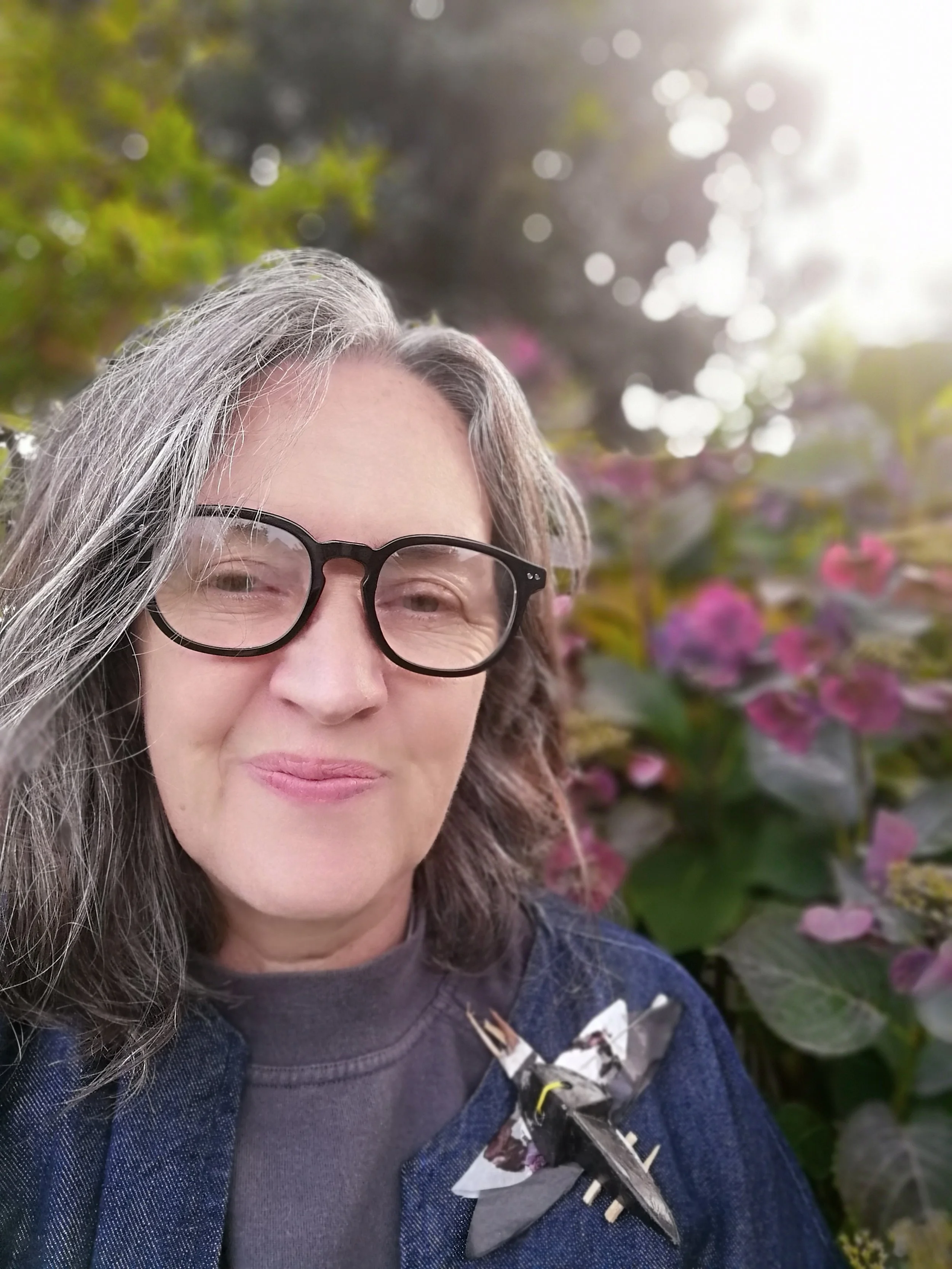 A woman with gray hair and glasses smiling outdoors, surrounded by pink flowers.
