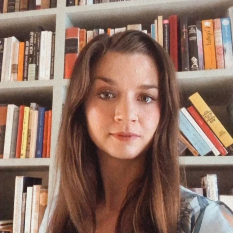 A young woman with long brown hair standing in front of a bookshelf filled with various books.