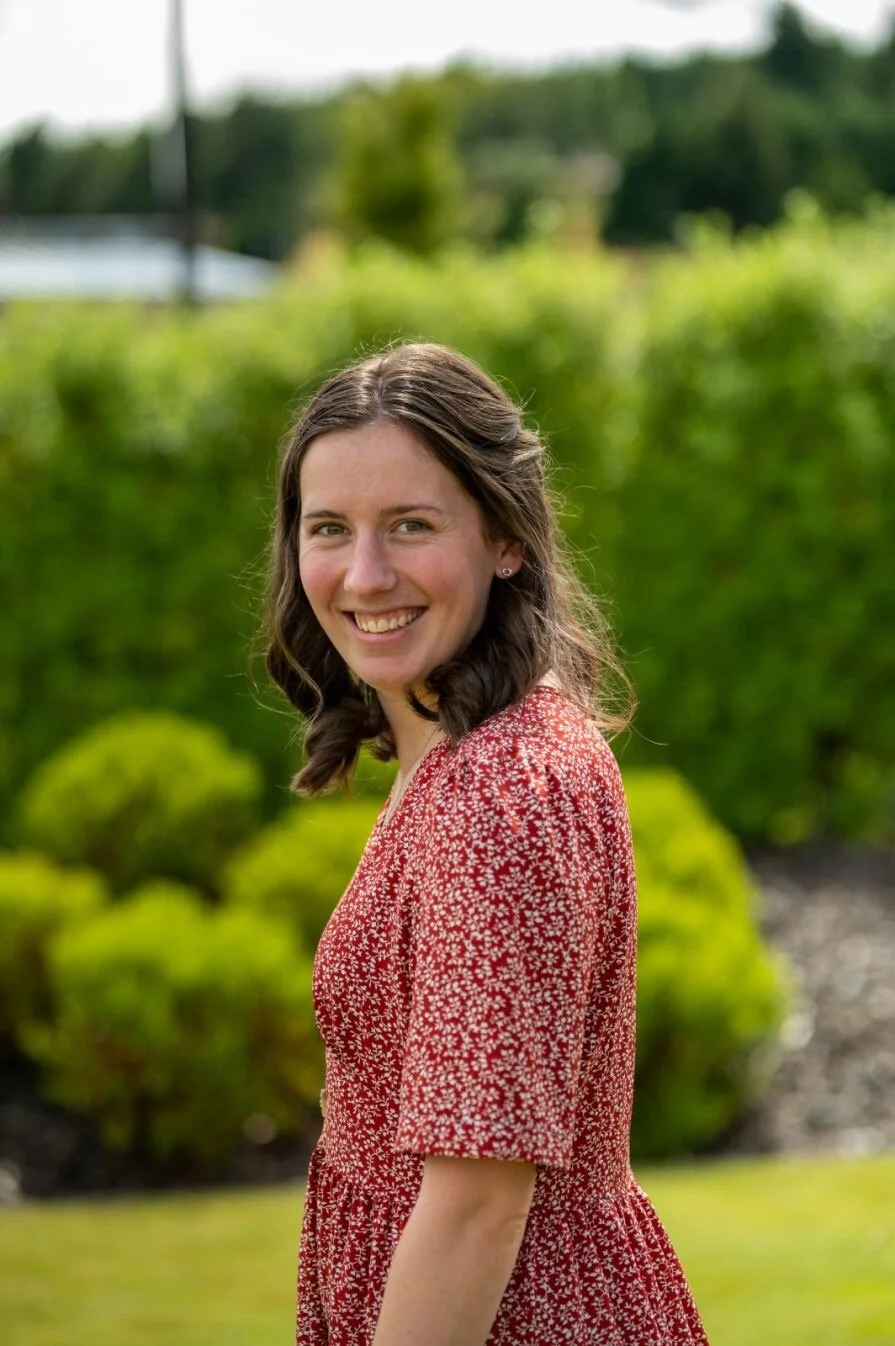 A young woman with shoulder-length brown hair, smiling, wearing a red dress with small white floral pattern, standing outdoors on a sunny day with greenery and bushes in the background.