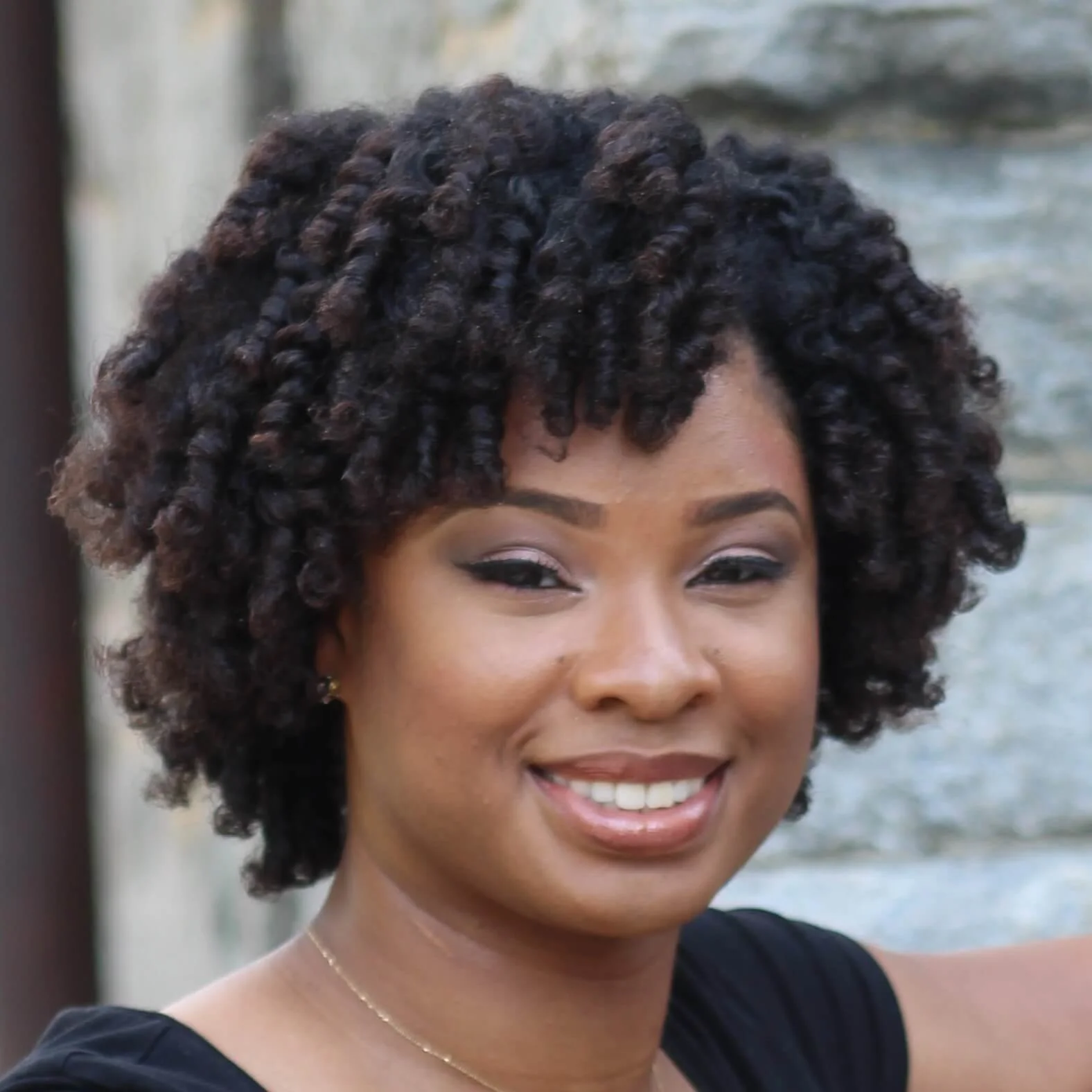 A woman with short, curly hair smiling outdoors with a stone wall in the background.