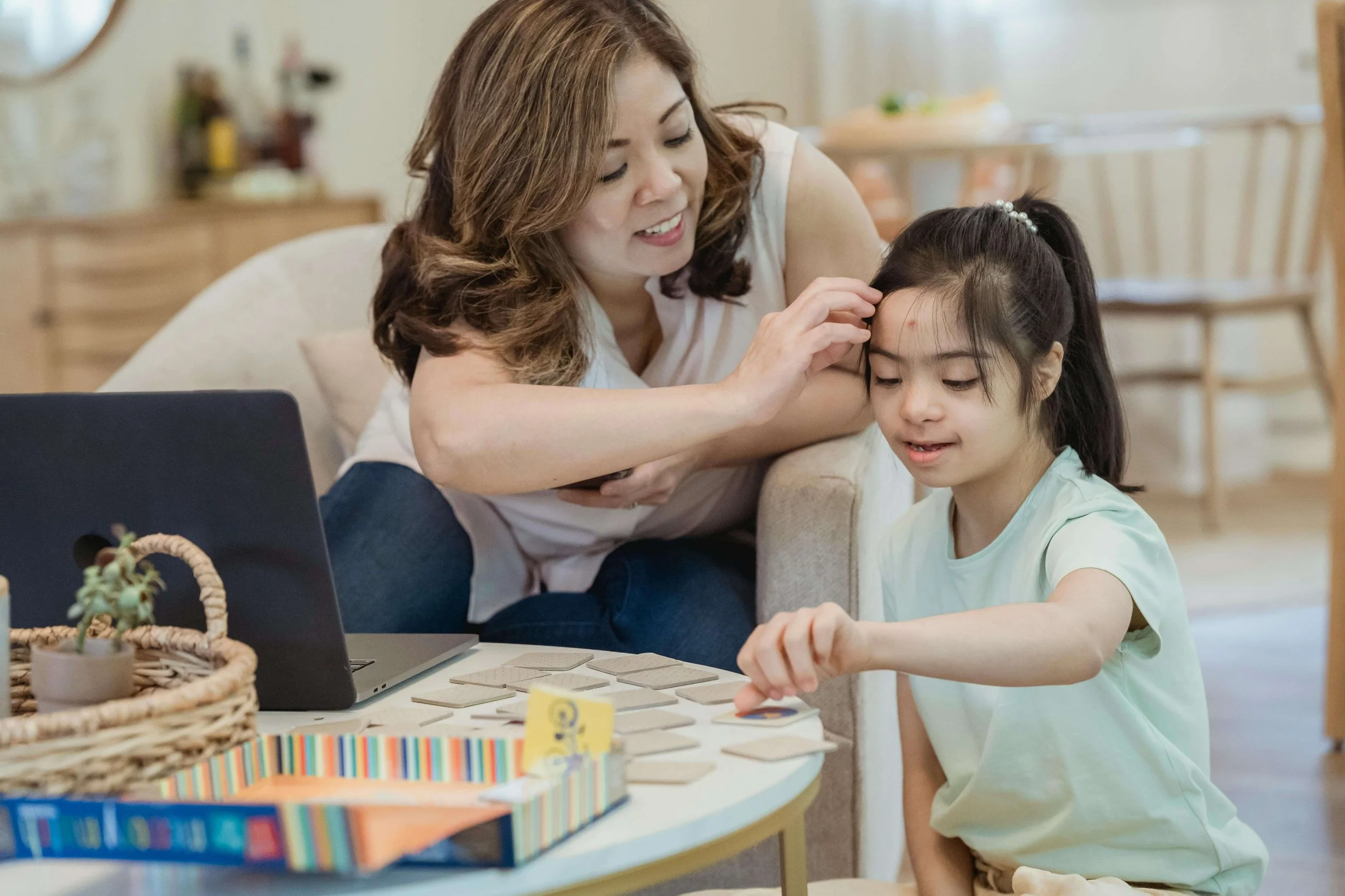 A woman is helping her daughter with a game on a table, touching the daughter's forehead while the daughter points at pieces on the table.