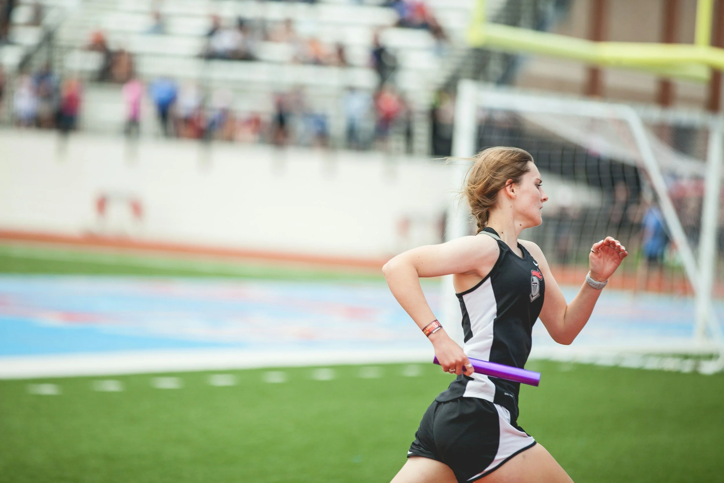 A female athlete runs on a track during a relay race, holding a purple baton in her hand.