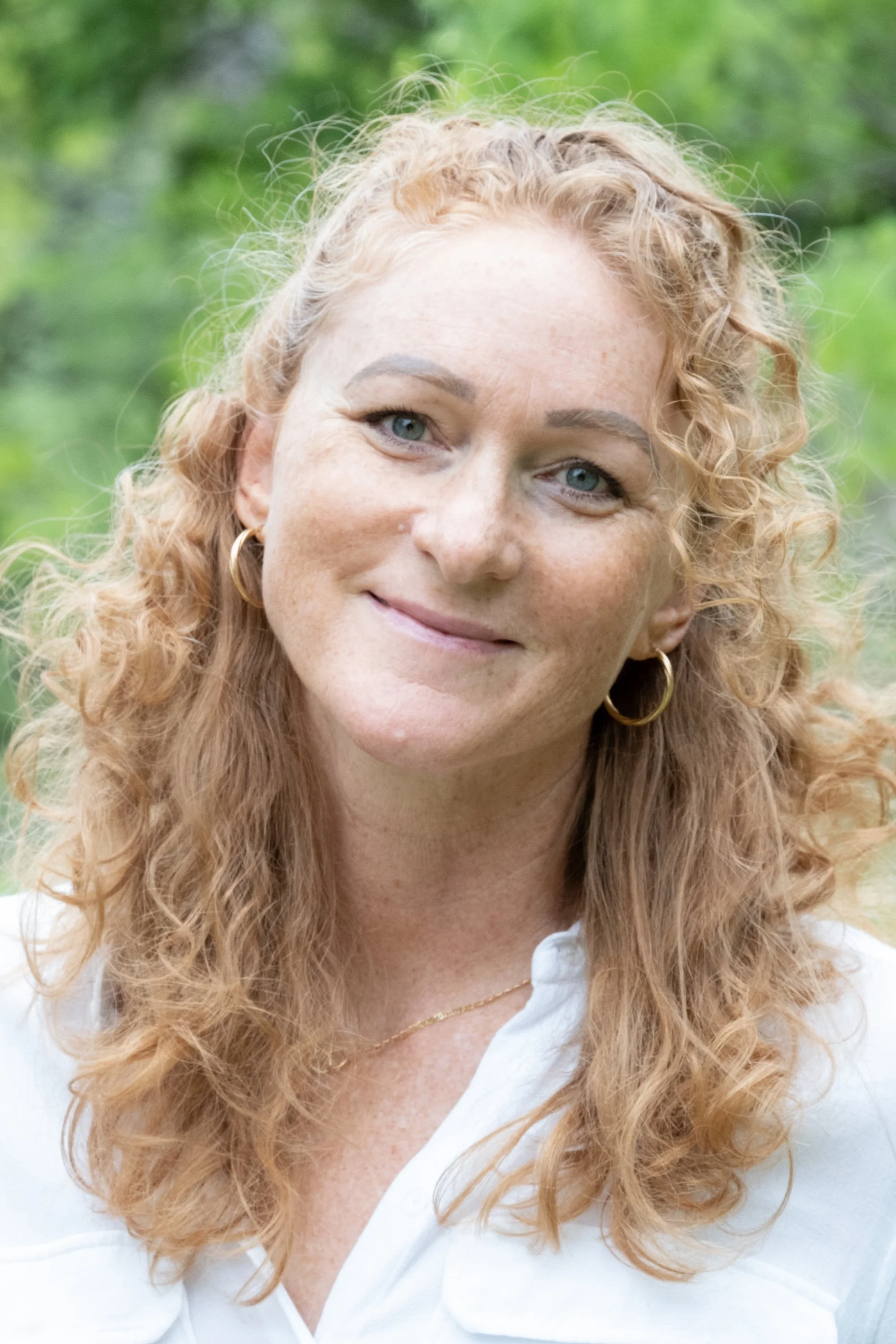 A portrait of Elli Overton, with curly red hair, blue eyes, and gold hoop earrings, smiling outdoors with green foliage in the background.