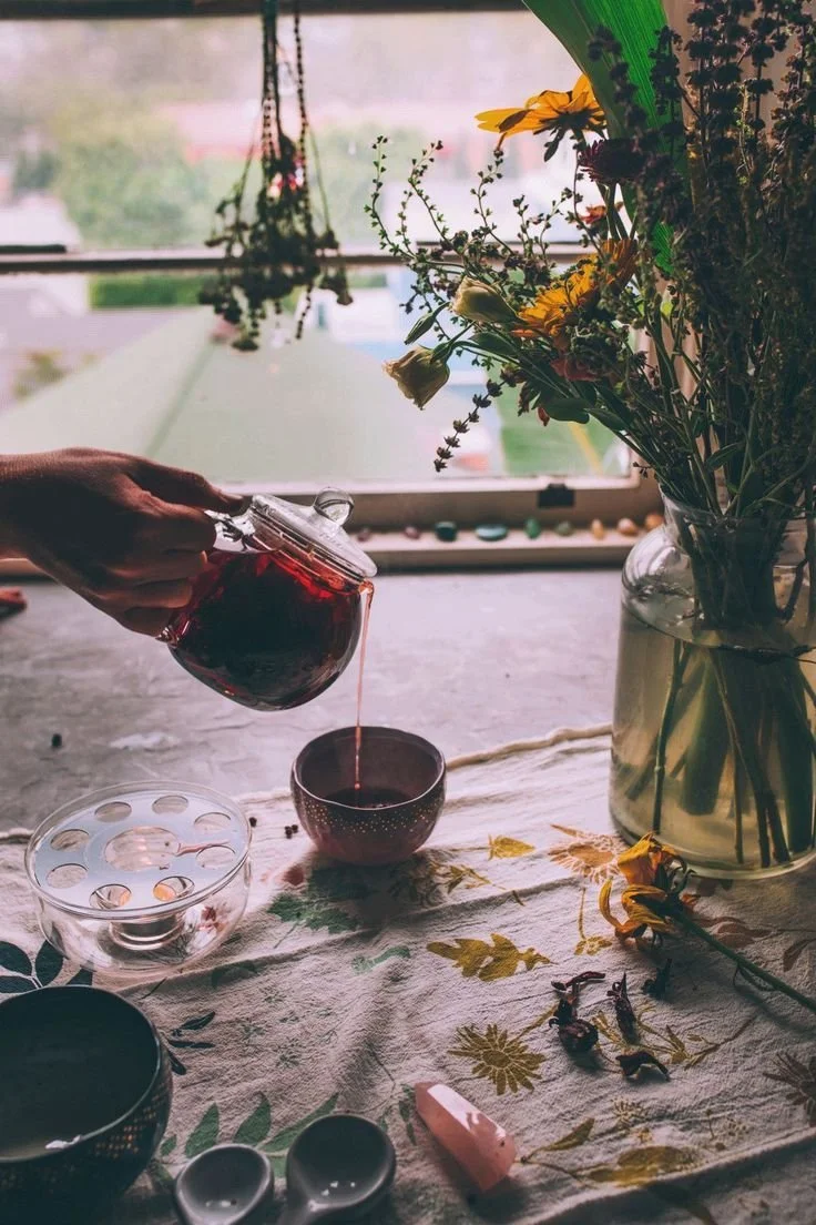 A hand pouring tea from a glass teapot into a cup on a table with a floral tablecloth. There is a large vase of flowers, decorative bowls, a pink crystal, and dried flowers on the table, with a window and greenery in the background.