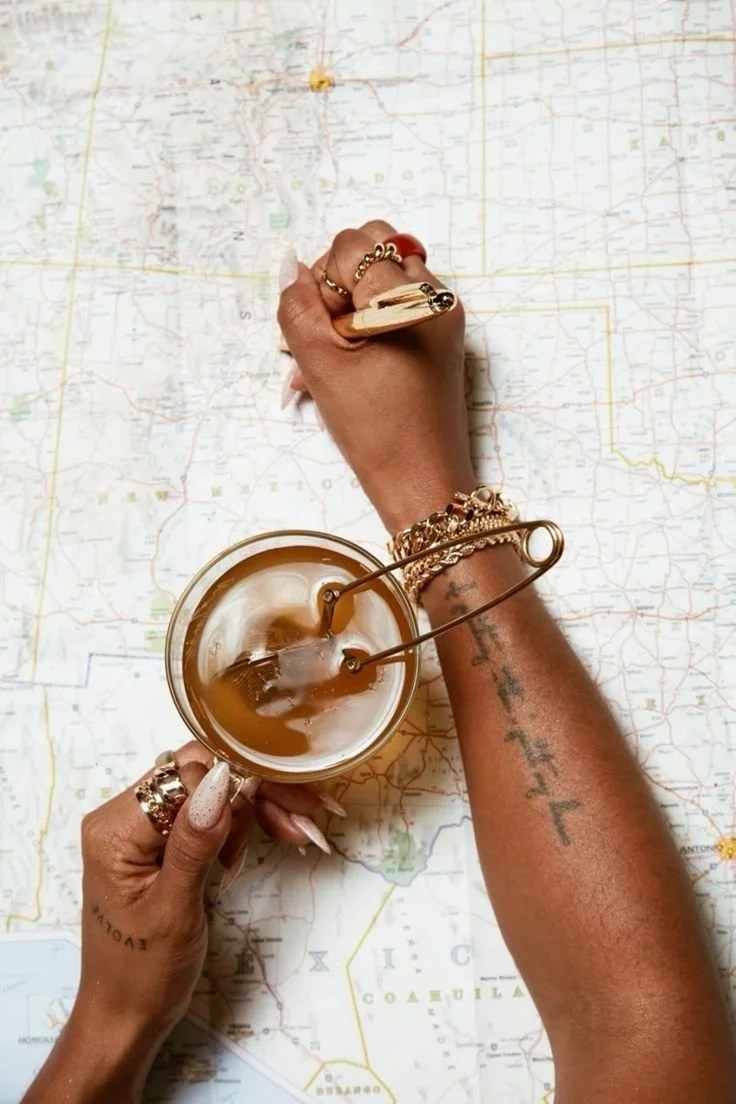Two hands, one with gold jewelry, are writing on a map with a gold pen. One hand holds a glass of iced coffee with ice and a straw. The map shows parts of Texas and nearby states.