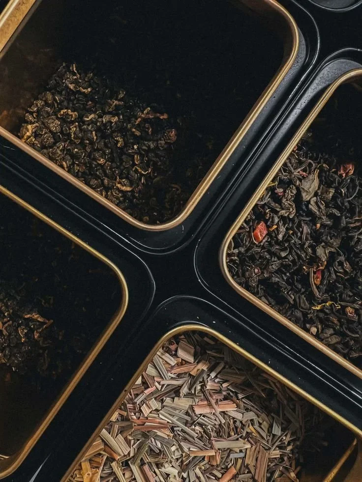 Four small containers filled with dried herbs or plant materials, including dark leaves, and shredded plant parts, possibly for tea or herbal blends.