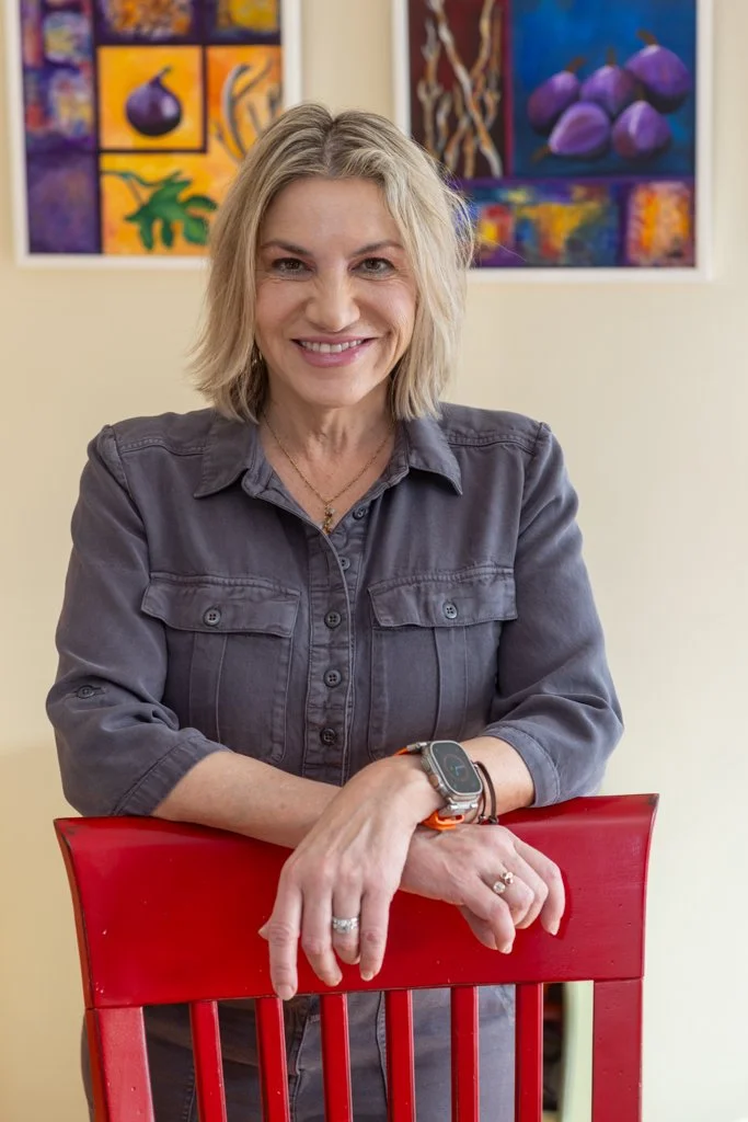 A smiling woman with shoulder-length blonde hair, wearing a gray button-up shirt and a smartwatch, leaning on the back of a red chair with colorful artwork hanging on the wall behind her.