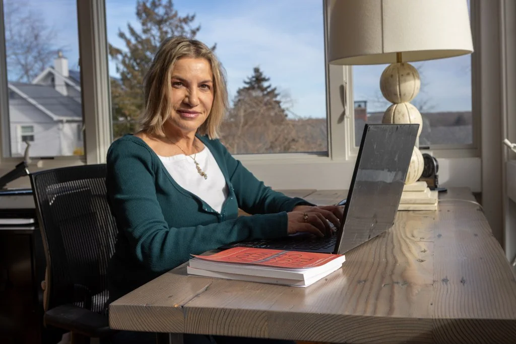 Woman sitting at a wooden desk working on a laptop with a large book beside her, near a window with a view of trees and houses outside.