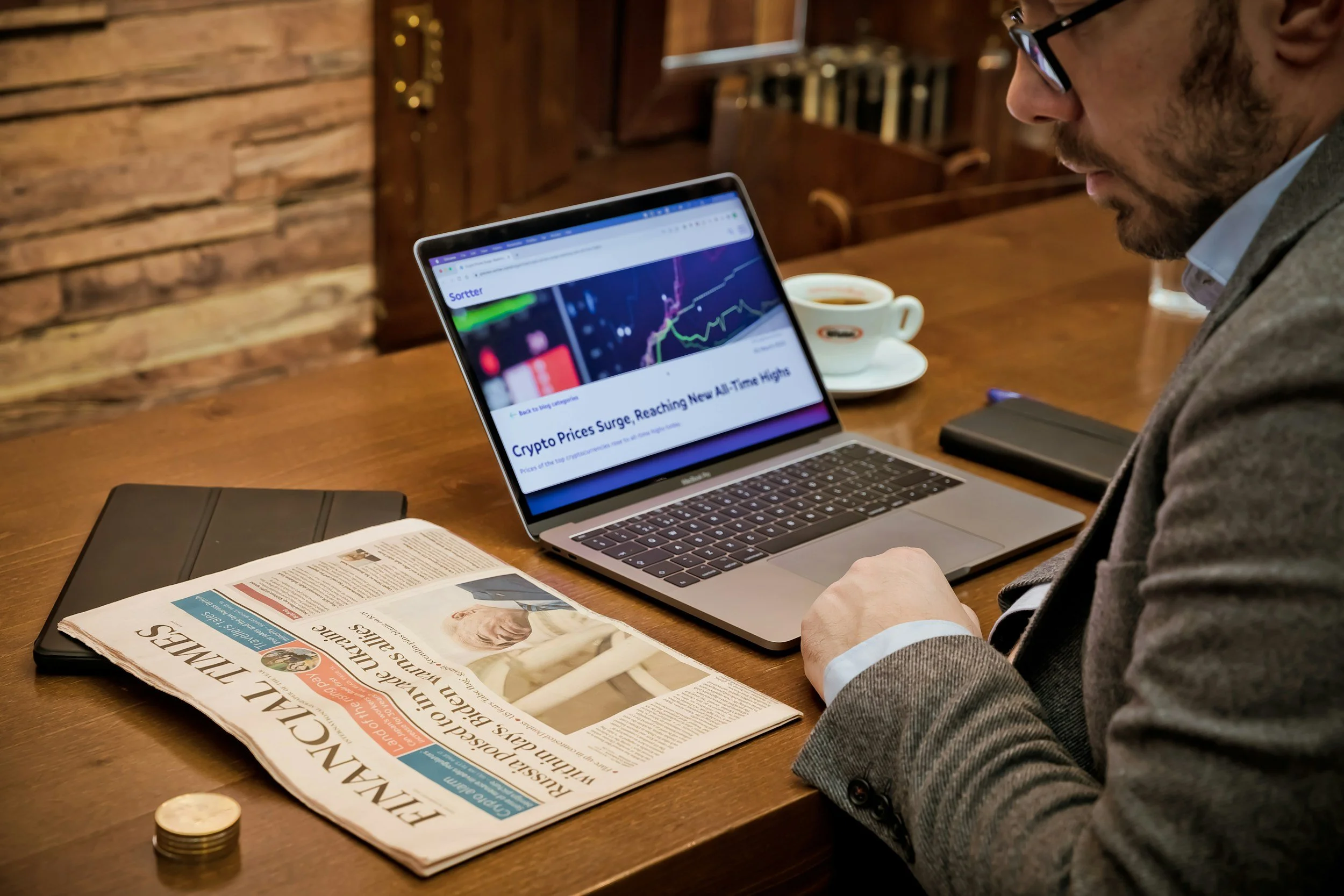 A man in a suit sitting at a wooden table with a newspaper, a laptop displaying a news article about crypto prices reaching an all-time high, a cup of coffee, a closed notebook, and a stack of coins.
