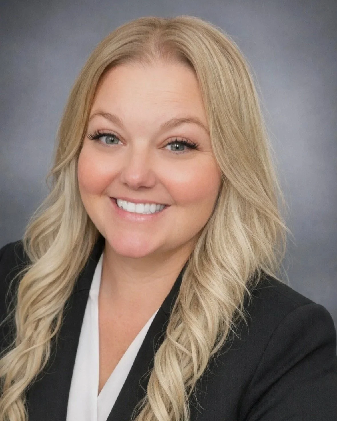 A woman with blonde hair styled in loose waves, smiling and wearing a black blazer over a white shirt, against a grey background.