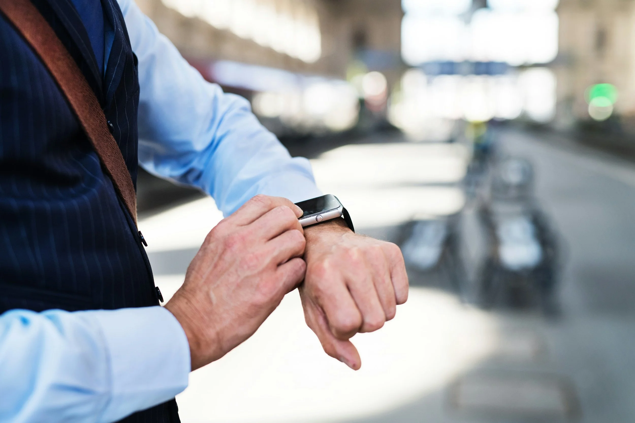 A person in a blue shirt and vest looking at a smartwatch on their wrist, while standing outdoors with a blurred city street background, including a motorcycle.