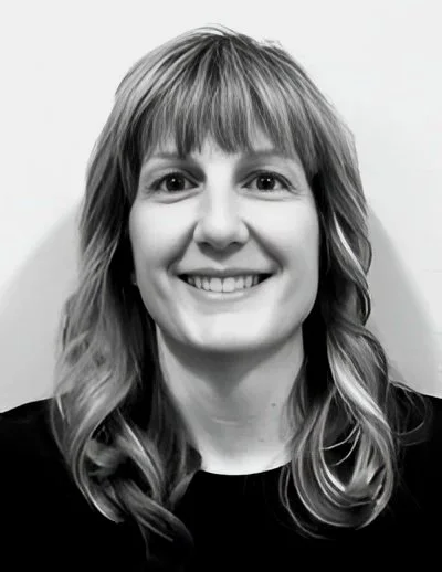 Black and white portrait of a smiling woman with shoulder-length wavy hair and bangs, wearing a dark top.