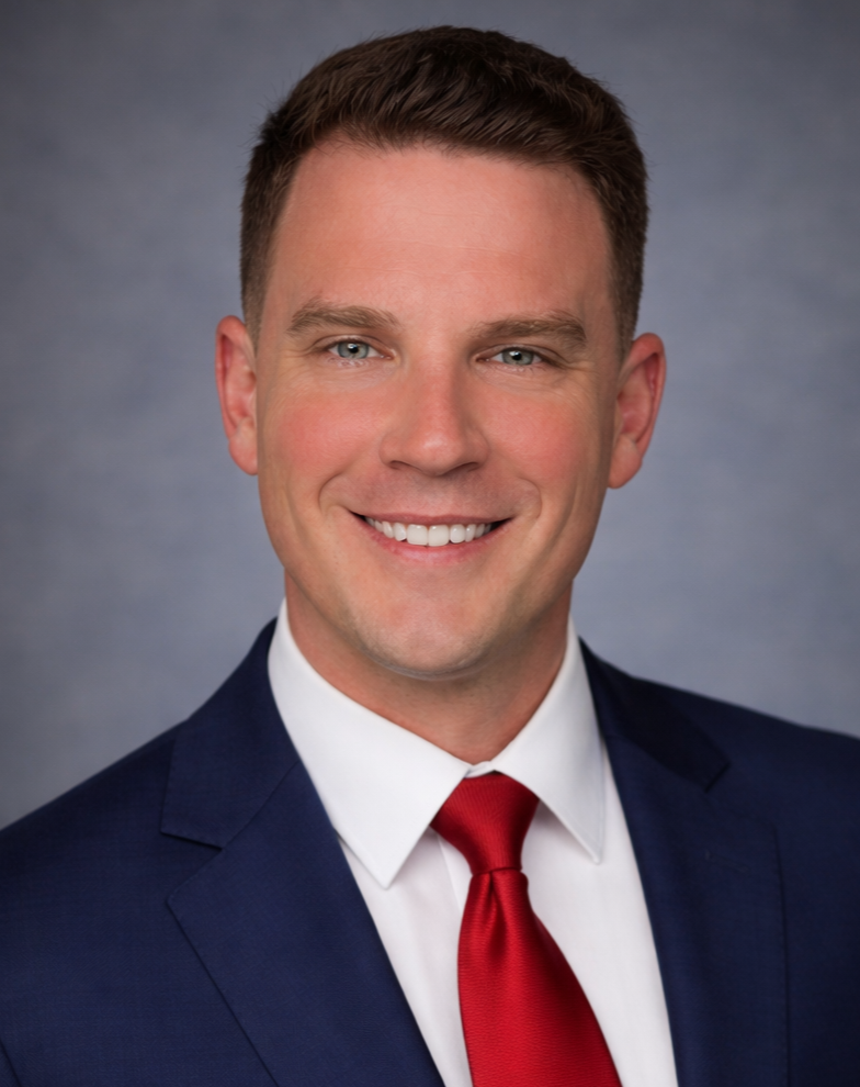 A professional headshot of a smiling man in a navy suit, white shirt, and red tie, against a gray background.