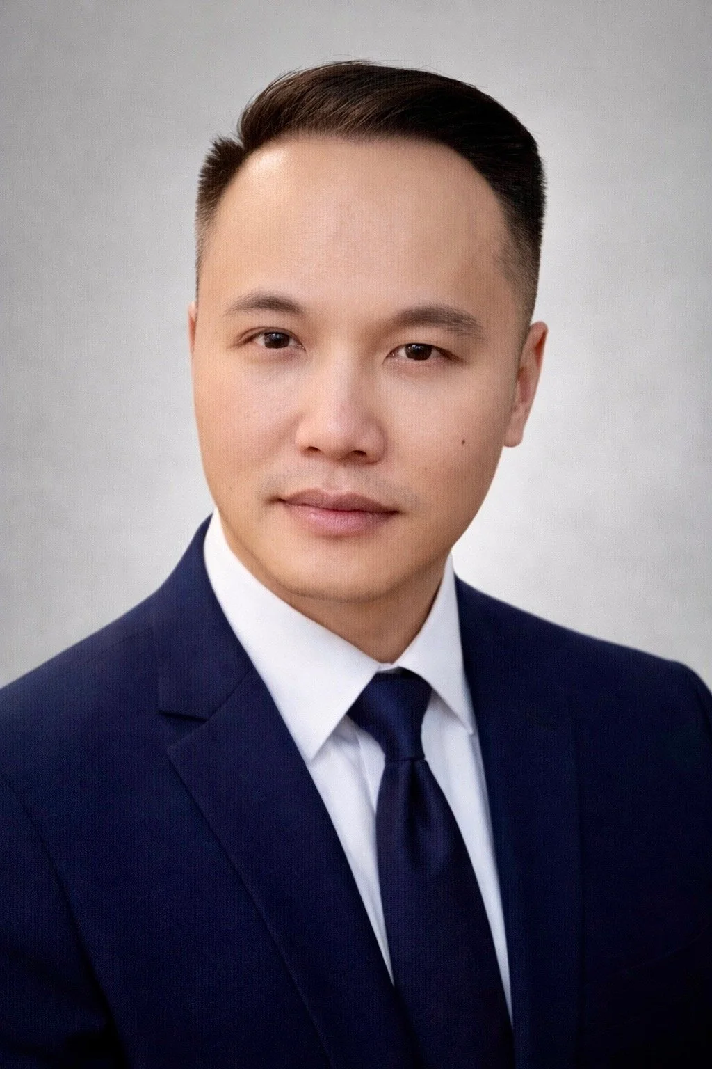 A professional headshot of a young Asian man in a dark suit, white shirt, and navy tie against a plain light grey background.