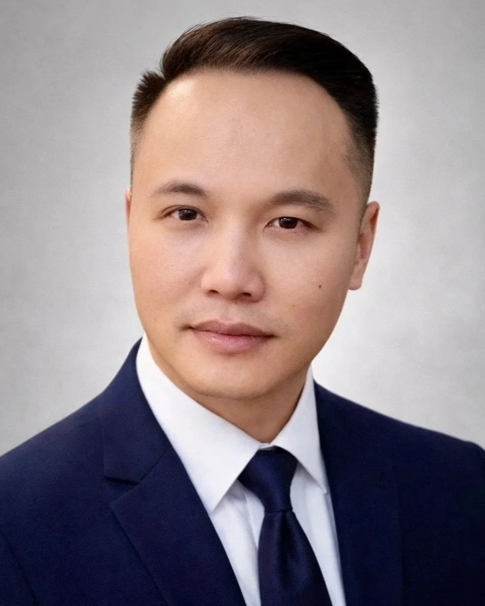A professionally dressed man in a dark suit and tie, with short dark hair, posing against a plain light background.