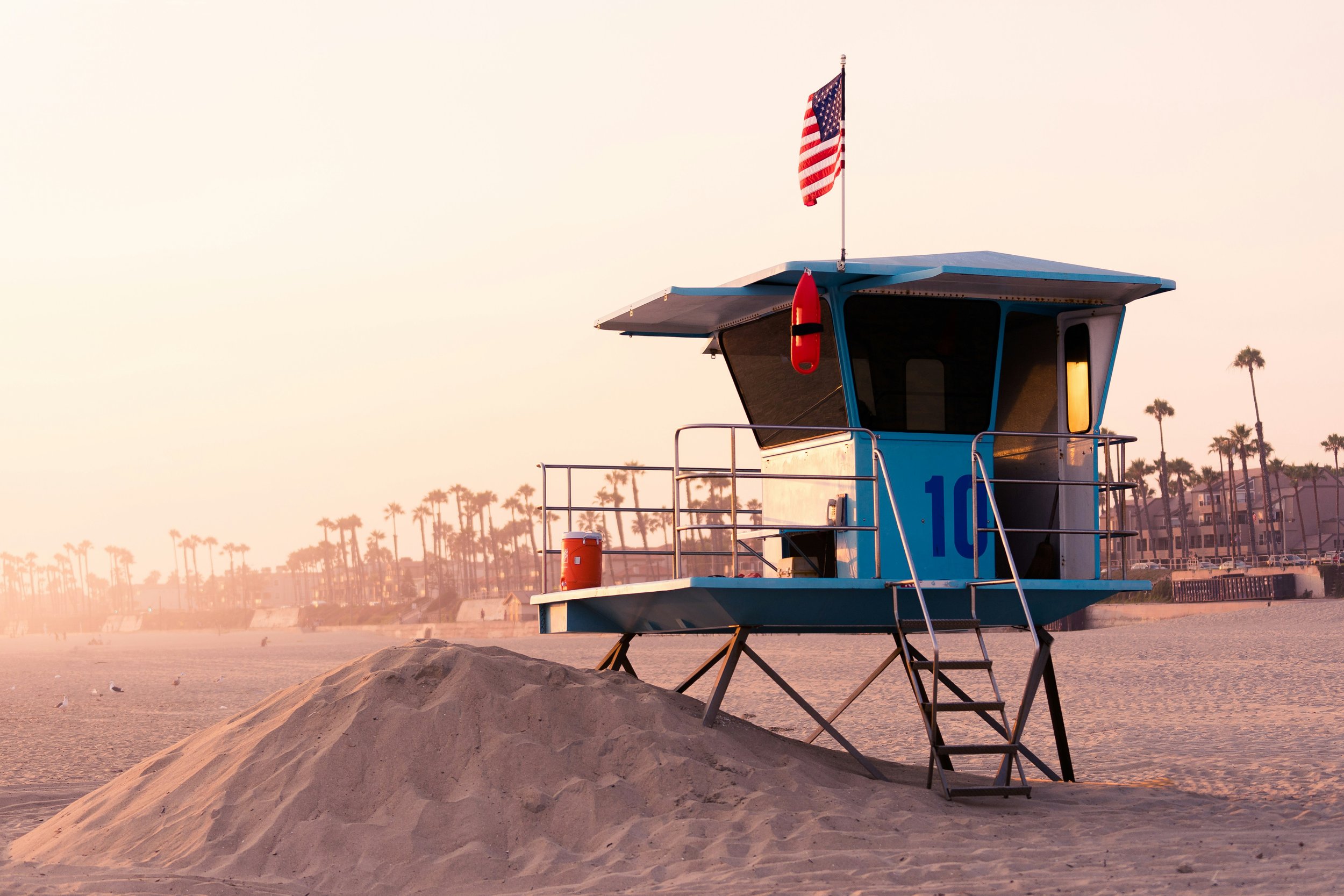 A lifeguard tower on a sandy beach at sunset, with an American flag, a rescue buoy, and a cooler, and palm trees in the background.
