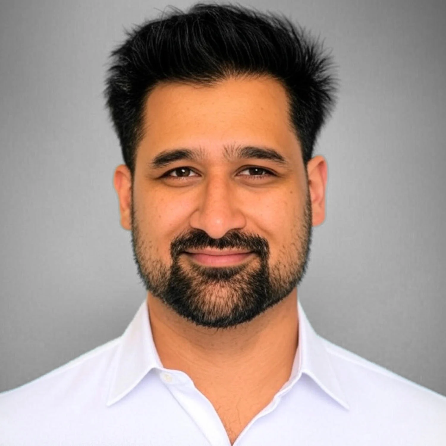 Headshot of a smiling man with dark hair and a beard, wearing a white shirt against a gray background.