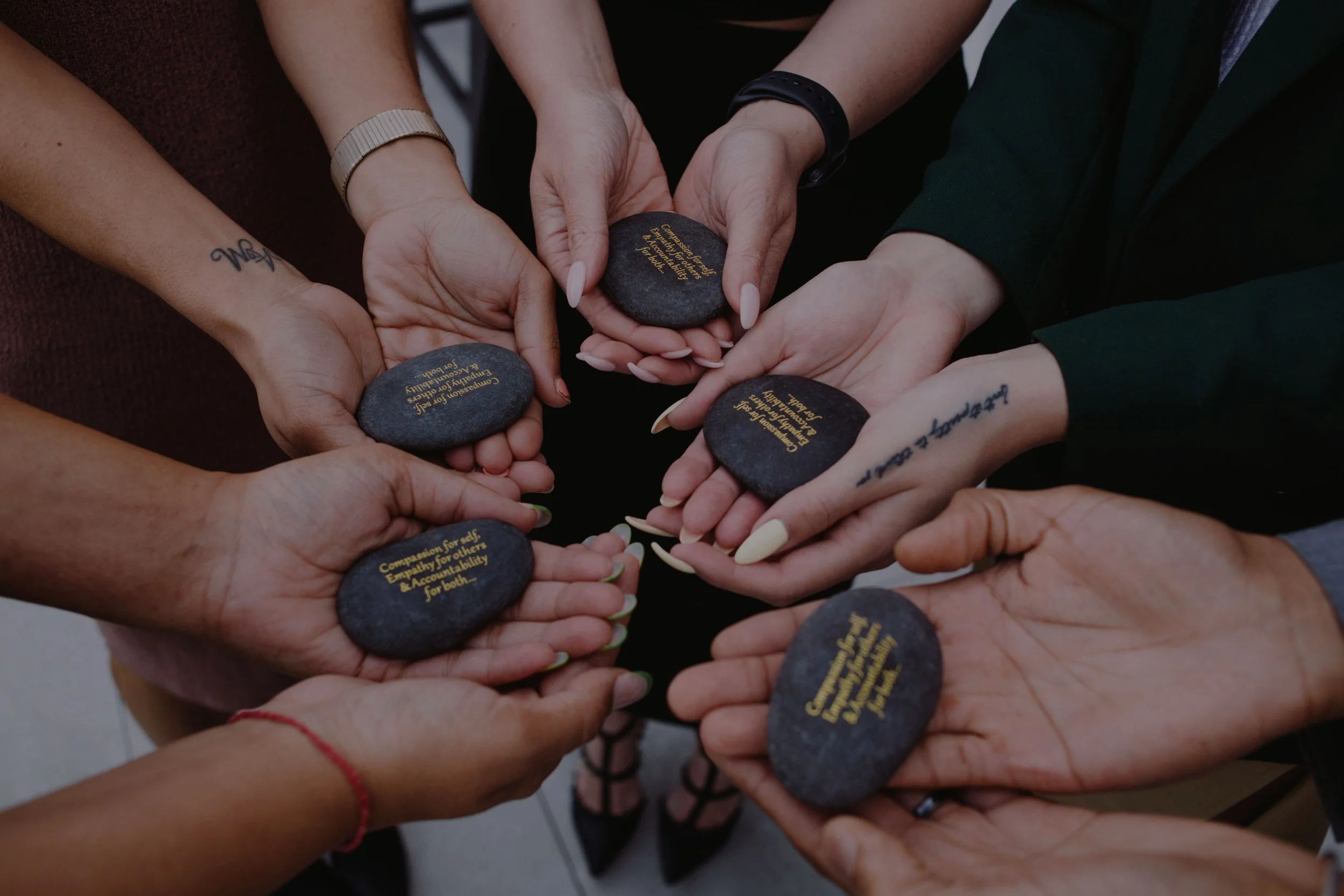 Many hands in a circle coming together in the center holding stones that say "compassion for self, empathy for others, accountability for both."