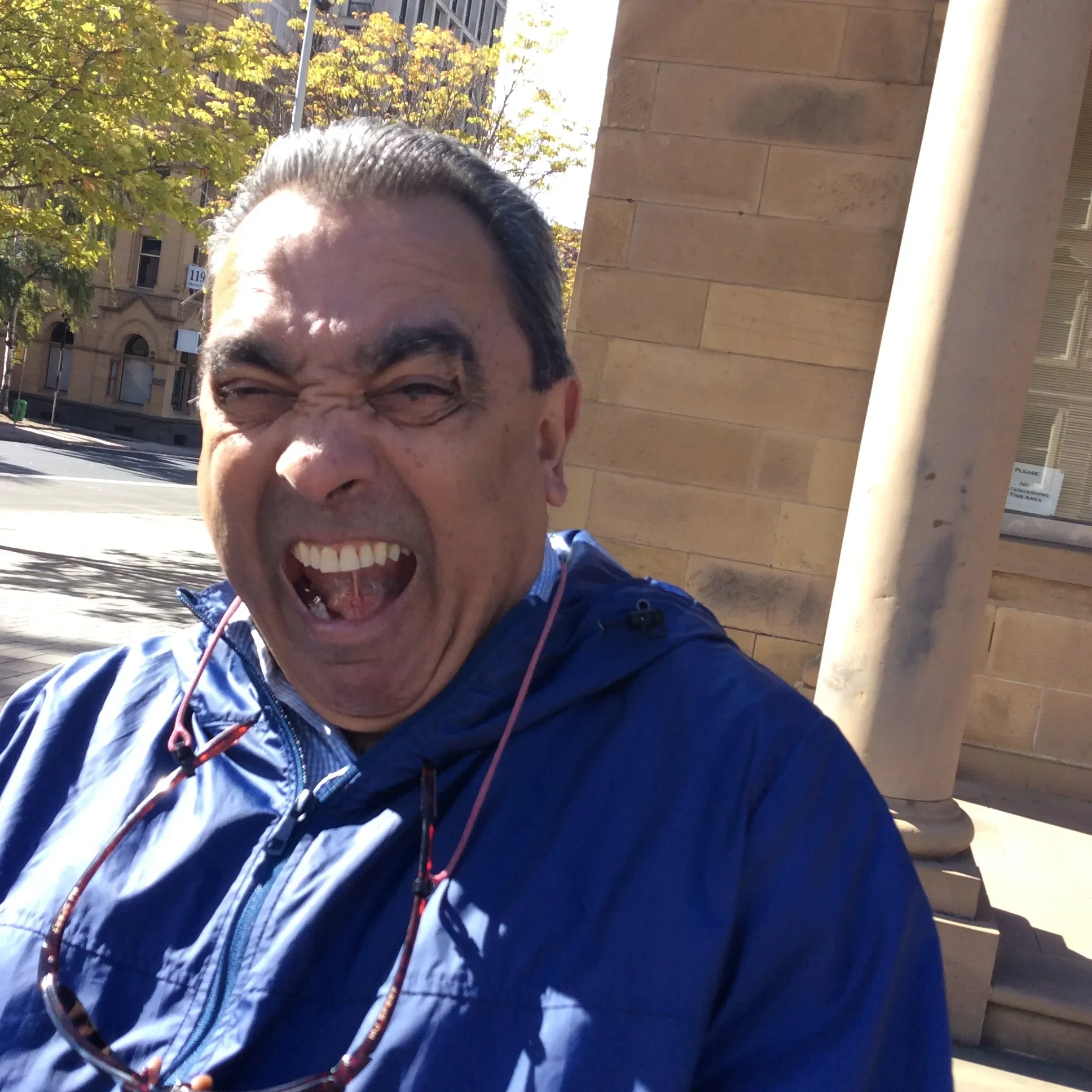 A man making a goofy face outdoors on a sunny day, wearing a blue jacket and sunglasses hanging from his neck, with trees and a building in the background.