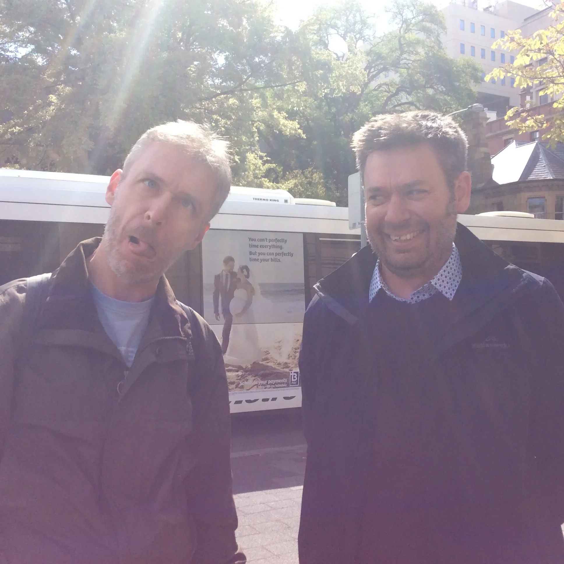 Two men standing outdoors on a sunny day, with a bus and trees in the background, one making a confused face and the other smiling.
