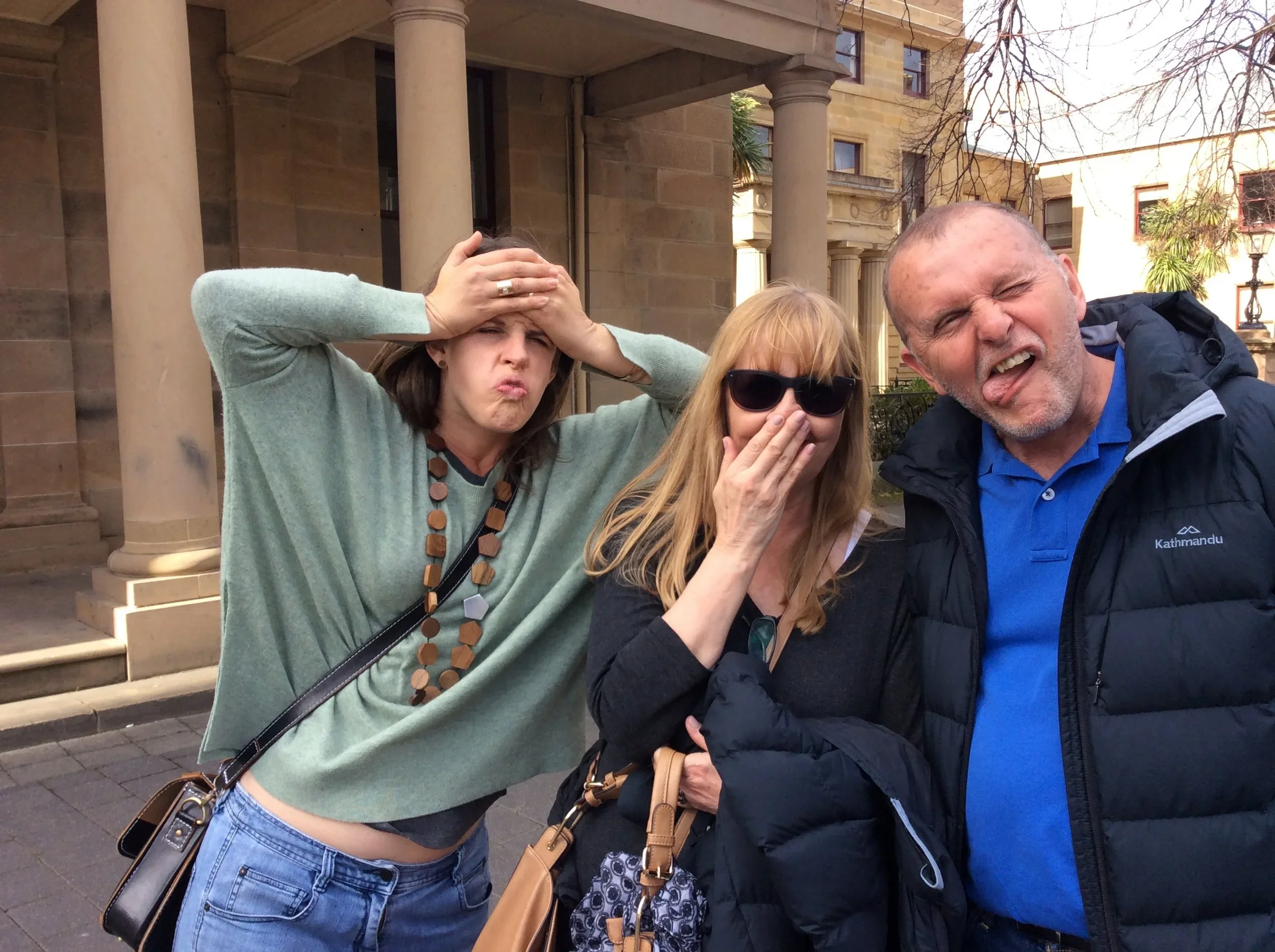 Three people making funny faces standing outside in front of a building with columns and trees.