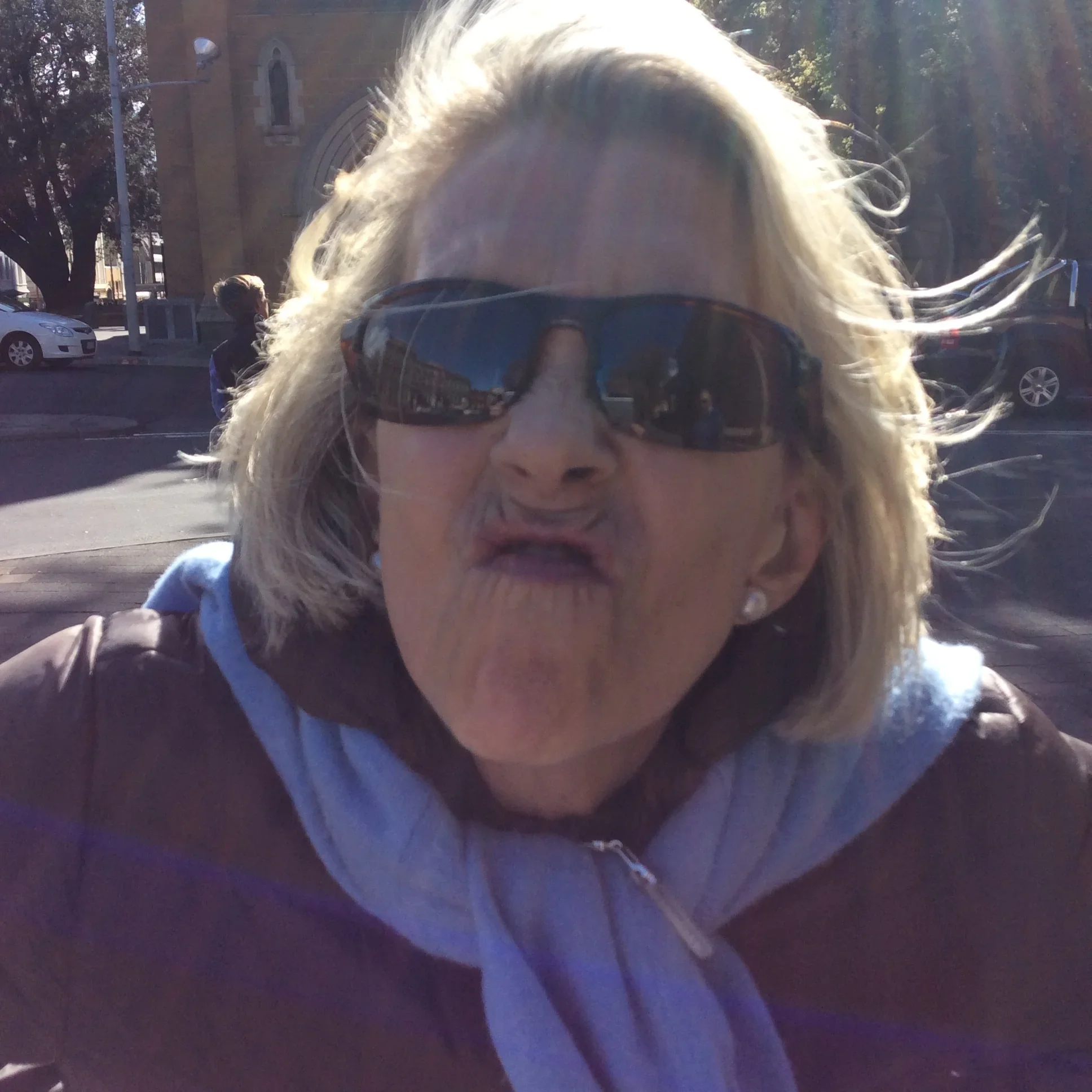A woman with blonde hair wearing sunglasses making a playful face, outdoors on a sunny day.