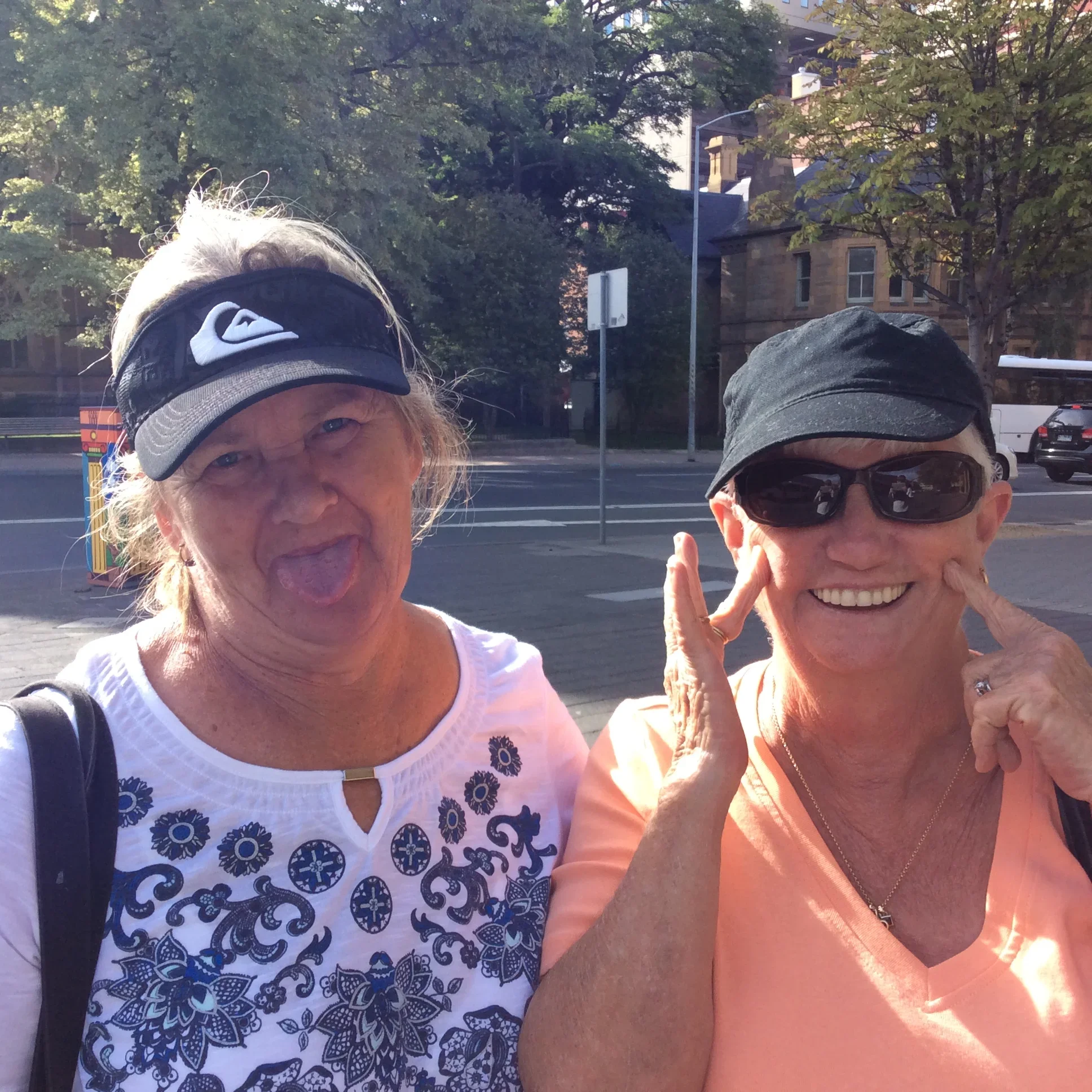 Two older women smiling and making playful faces outdoors in sunlight, wearing sunglasses and hats on a city street.
